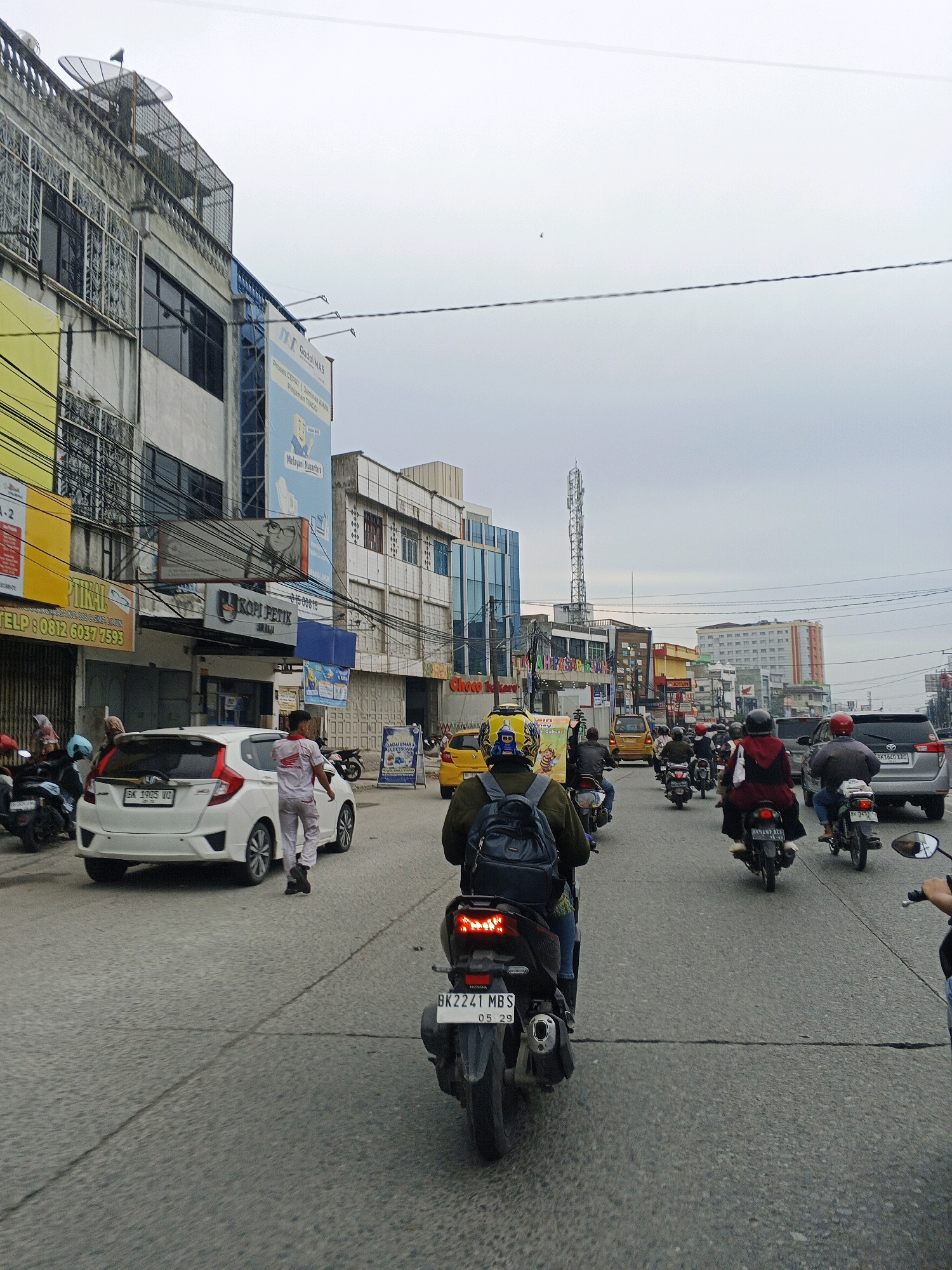 The image depicts a street scene in the city of Medan, Indonesia, likely on a cloudy day. The perspective is from a vehicle driving on a multi-lane road filled with traffic, predominantly motorcycles. On the left side of the road, there is a row of buildings with various signs and advertisements. One building has a large yellow sign with black text, and another has a blue sign with an illustration of a person. Several power lines are visible crisscrossing the sky.
In the foreground, a white car is parked on the left side of the road, with its rear facing the camera. A person in light-colored uniform, possibly a worker, is standing next to the car. Several motorcycles are also present in the scene. In the mid-ground and background, more vehicles, including cars and motorcycles, are navigating the road. Buildings of varying heights and architectural styles line the street. A tall communication tower is visible in the distance. The road surface appears to be asphalt, with some visible cracks and markings. The overall atmosphere suggests a busy urban environment with ongoing traffic.