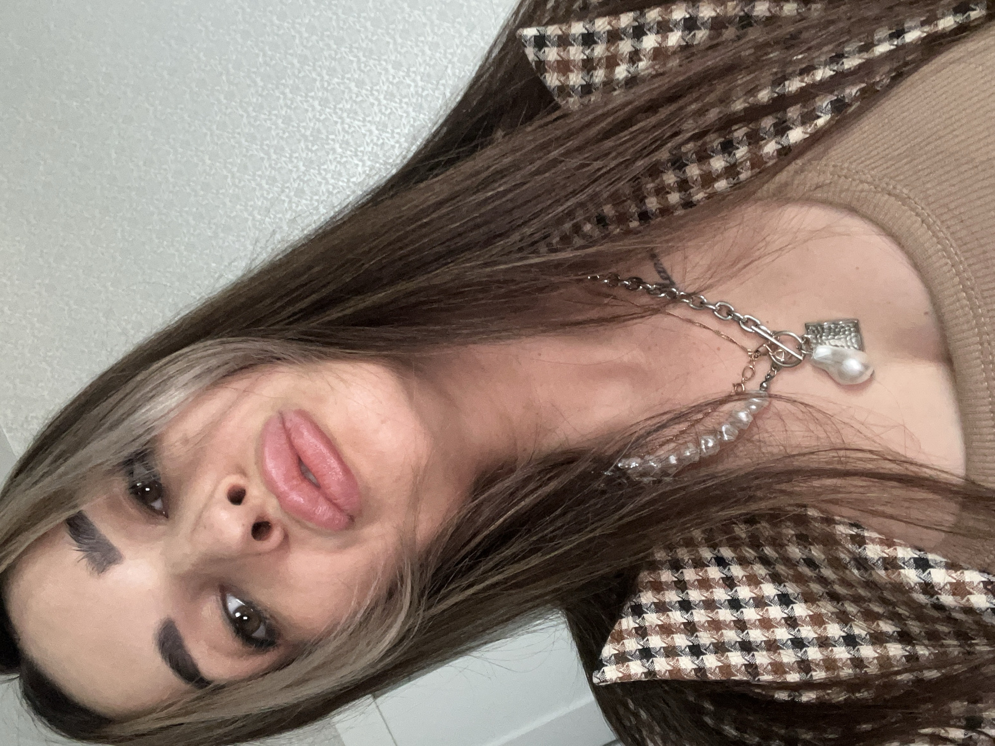 An adult female is depicted in an upward-angled close-up selfie, occupying the central and upper portions of the frame. She possesses long, straight, dark brown hair with blonde highlights framing her face, parted centrally. Her eyebrows are dark and defined, and her brown eyes gaze directly at the camera. Her lips are visibly full, colored with a light pink pigment, and are pursed in a distinct pout. She is attired in a light brown, ribbed inner top, beneath a blazer featuring a brown, black, and cream-colored houndstooth or checkered pattern. Multiple necklaces are worn: a choker composed of irregular pearls, a thick silver-toned chain necklace with a T-bar clasp from which a large baroque pearl and a smaller textured rectangular silver-toned pendant are suspended, and a subtle gold-toned chain beneath the other layers. The background is a light gray or off-white wall exhibiting a fine, granular texture, indicating an indoor environment. The precise location within the city of Unknown, Unknown, is indeterminate based on the visual information provided.