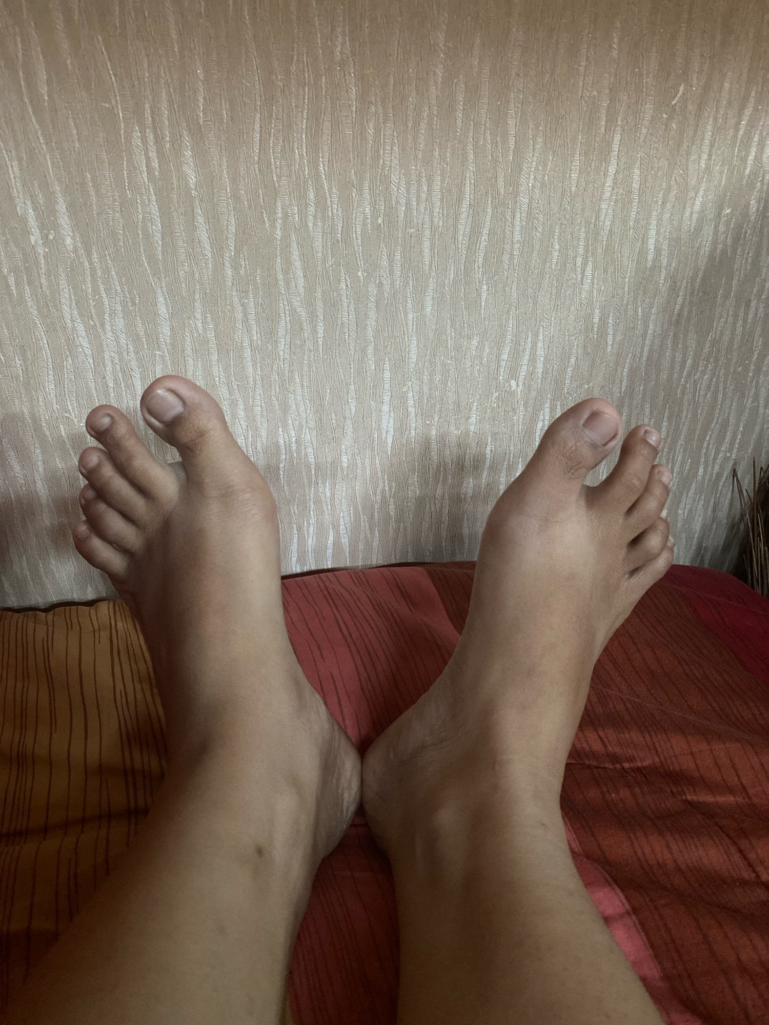 The image shows a close-up, eye-level view of two bare feet resting on a patterned surface. The feet are positioned with the toes pointing upwards and slightly outward, forming a V-shape. The skin tone of the feet appears to be medium to dark. The background consists of a textured, light-colored wall. The surface beneath the feet has a reddish-orange hue with subtle linear patterns. The overall lighting suggests an indoor setting. There is no indication of the location being in Jimbaran Kulon, Indonesia.