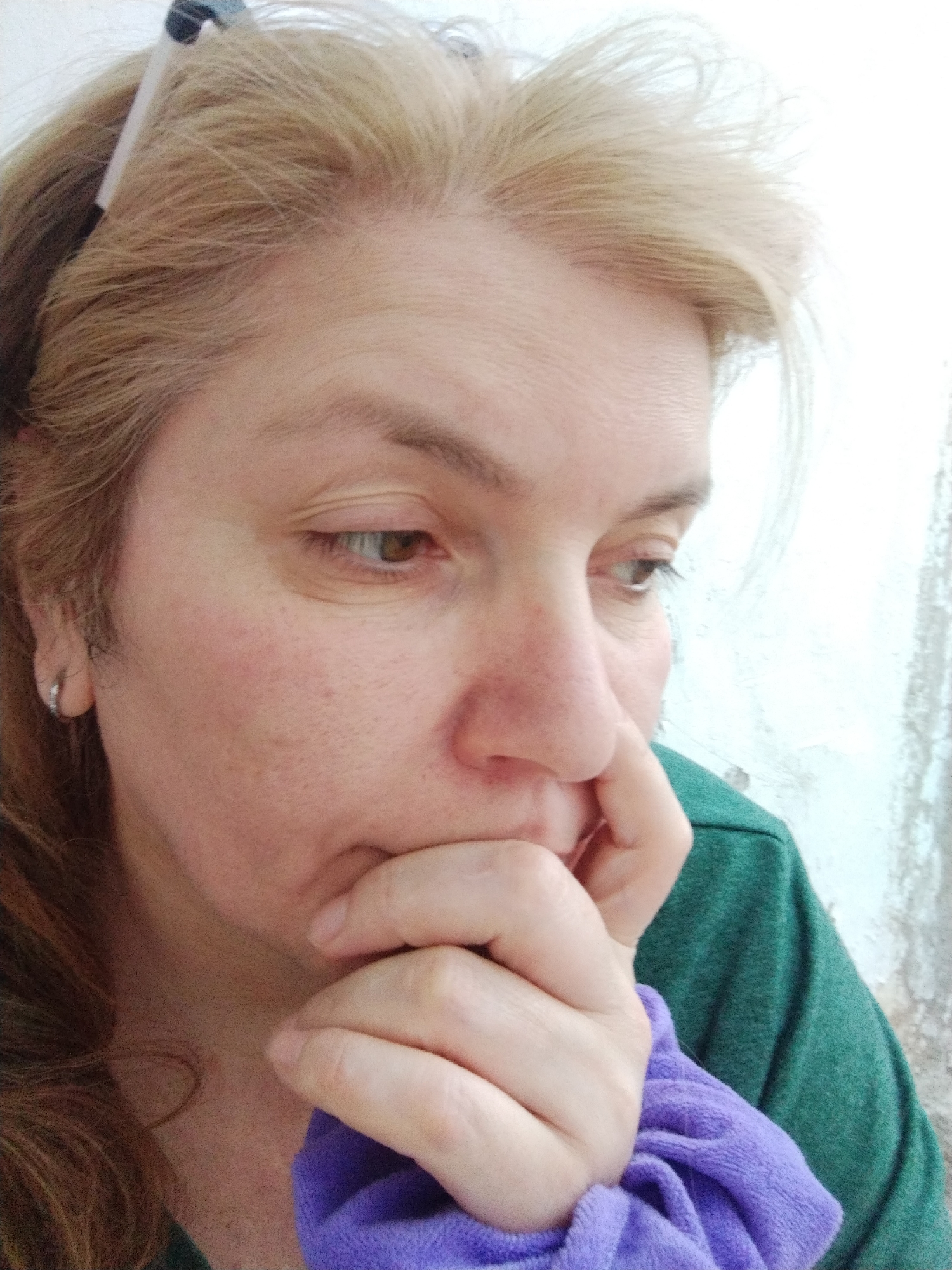 A middle-aged woman with fair skin and blonde hair is the primary subject of the image. She is wearing a green shirt and is holding a purple fabric item near her face. Her expression appears thoughtful or concerned. She is wearing a small hoop earring on her left ear. A hair clip is visible in her hair. The background features a plain, possibly textured wall, and it is difficult to determine the exact location. The image does not provide enough information to confirm the location in Al Mas'oudi, United Arab Emirates.