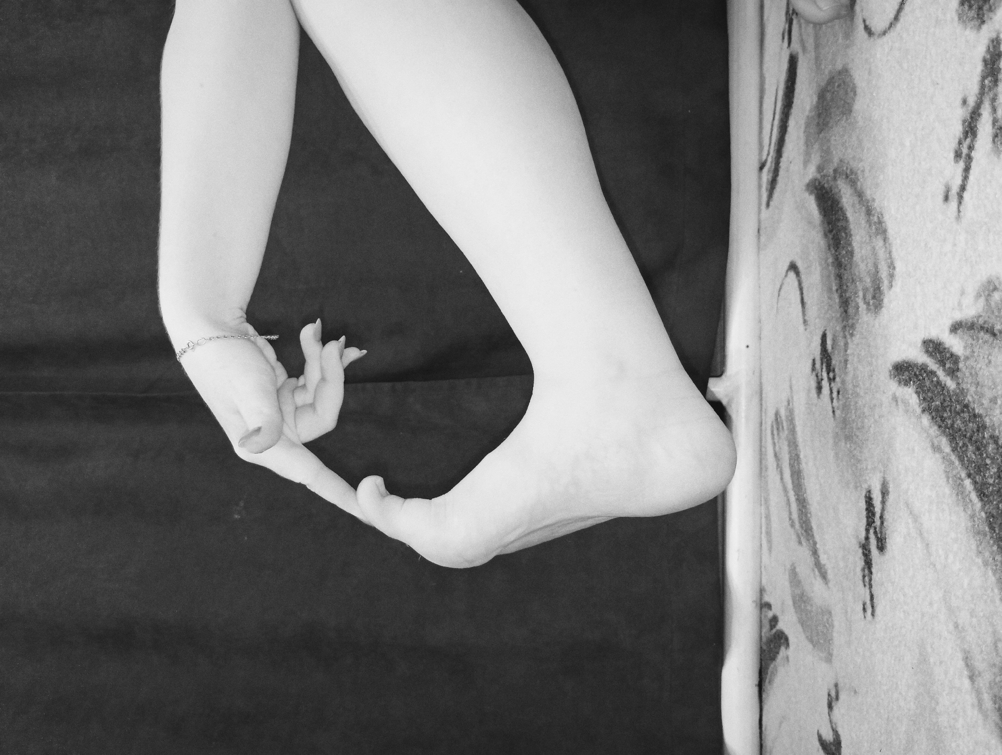 A black and white photograph depicts a pale forearm and hand holding a foot. The forearm is positioned on the left side of the frame, and the hand is bent backward, with the fingers touching the big toe. The foot is at the right side of the frame. The background is a dark, flat surface, with a patterned surface visible on the right side. The location is not identifiable.