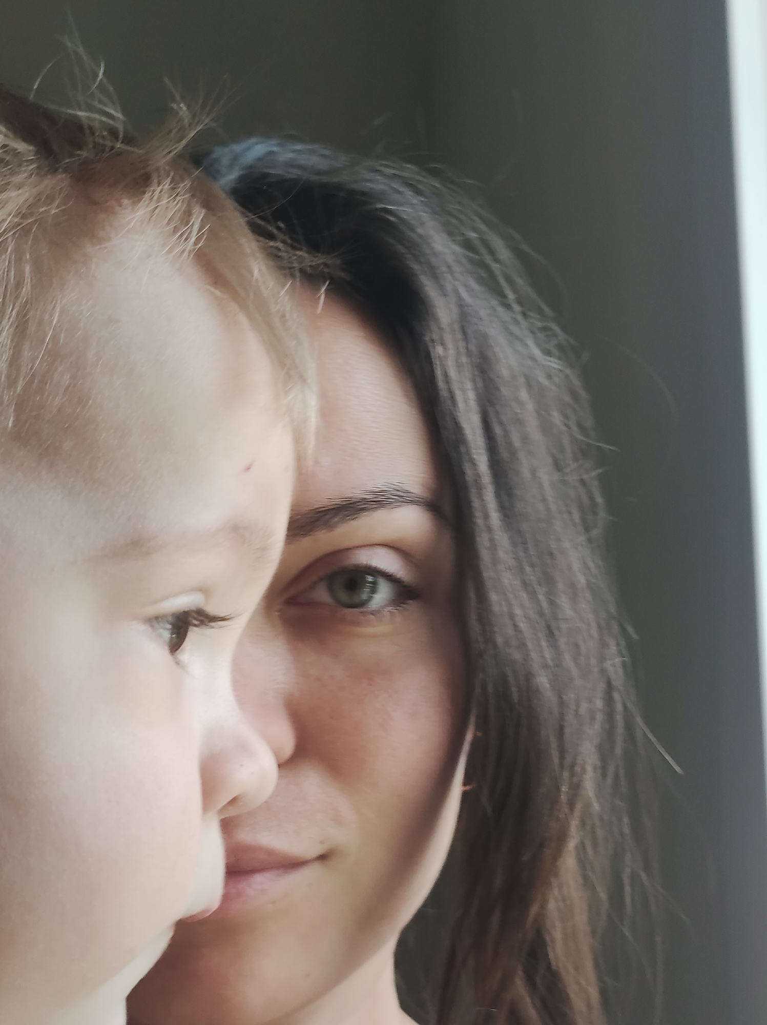 The image presents a close-up of a woman and a child. The woman has dark hair and a visible eye. The child, situated to the left, has blond hair and a visible eye. Both faces are partially visible, creating a sense of intimacy. The background is neutral in tone. The location is Unknown, Unknown.
