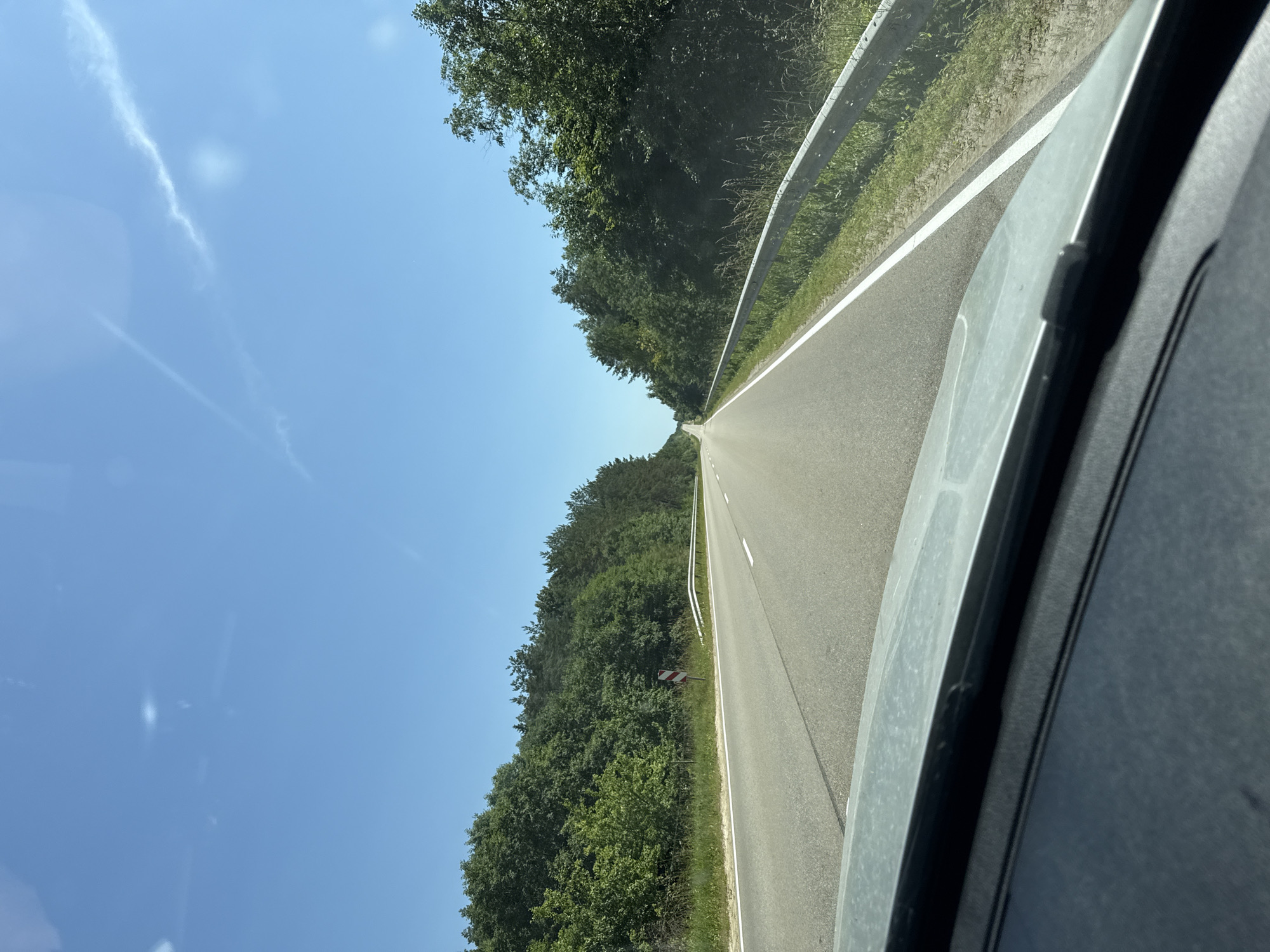 The image captures a view from inside a vehicle traveling on a paved road in Subate, Latvia. The road curves to the right and is bordered by lush green trees on both sides. A metal guardrail runs along the right side of the road. The sky is a clear, bright blue with a few wispy clouds. The foreground shows a portion of the vehicle's dashboard. The scene suggests a sunny day on a highway, likely in a rural or forested area outside the main city.