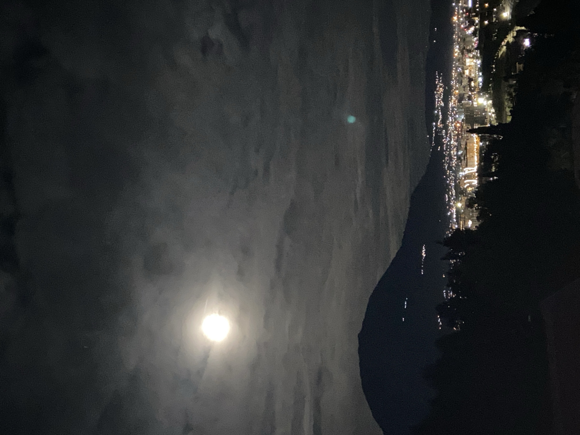 This image is a night-time shot of the moon, a mountain, and a city. The moon is a bright, round orb near the bottom left of the frame, partially obscured by clouds. A dark mountain range or hill is positioned to the right of the moon, separating it from the city lights. The city lights are densely clustered at the top right of the image, indicating an urban area. The scene appears to be in Šušanj, Montenegro, due to the coastal cityscape at the right of the mountain.