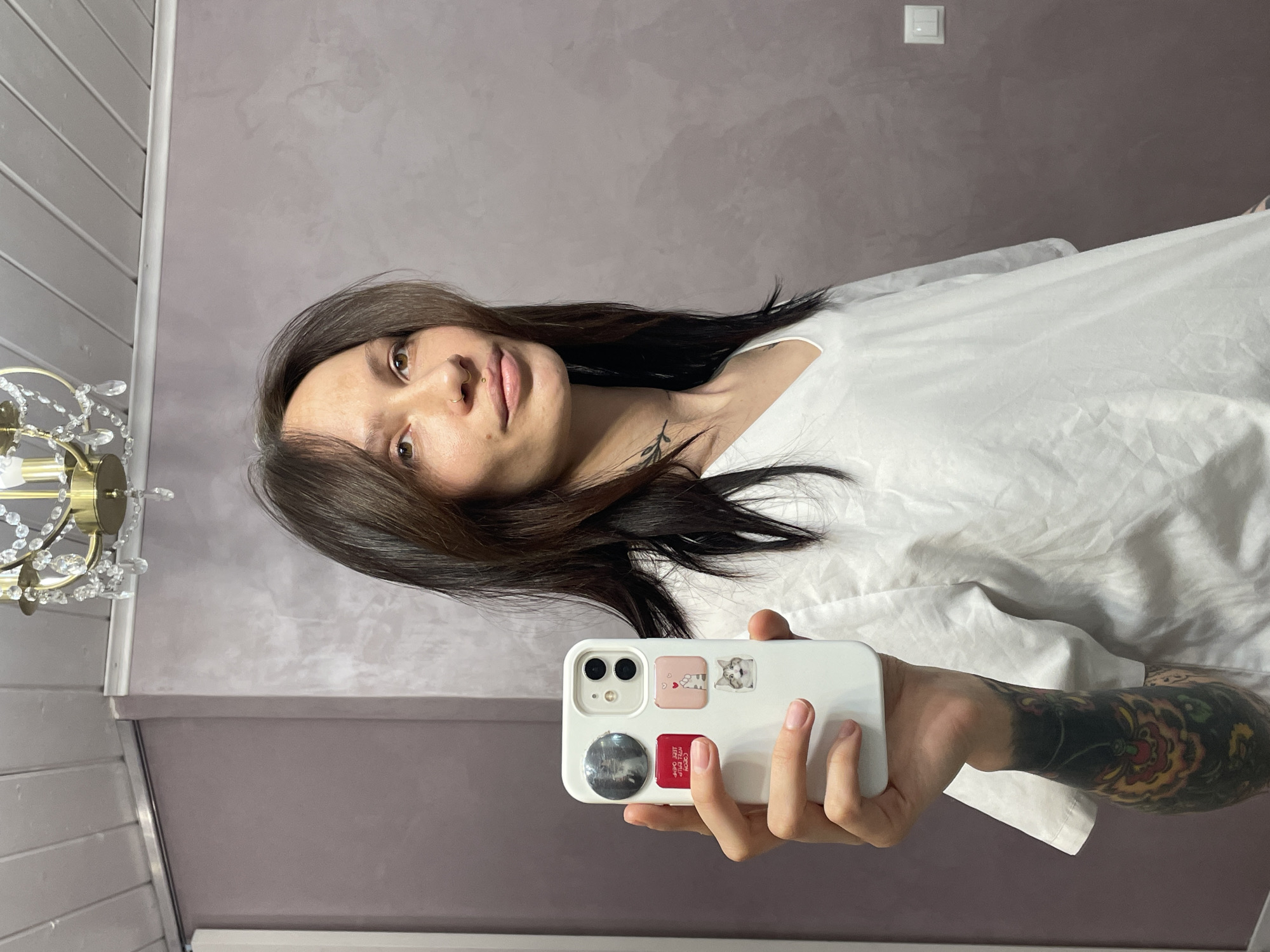 The image displays a woman taking a self-portrait indoors, holding a white smartphone in her right hand. She is angled across the frame, with her head towards the upper left. She has long, dark brown hair, brown eyes, and fair skin, with a small gold nose ring on her right nostril. Her expression is neutral with a slight smile. She wears a loose white shirt. Her left arm is visible from the elbow down, covered by a vibrant full-sleeve tattoo featuring colorful floral or ornate patterns in red, yellow, green, and dark shades.
The white smartphone case is adorned with several stickers: a small pink square with a white heart and tiny text, a grey sticker with a white cat's head, a red rectangular sticker displaying "CROWN," "LITE CORD," and possibly "SIZE CROP" in white text, and a round, dark-colored pop socket-style sticker with an image resembling a moon or a planet.
The background consists of a textured greyish-purple wall. To the far left, a small, ornate gold-colored chandelier with clear crystal drops is partially visible. White horizontal panels are seen in the upper left corner, and a white electrical switch is mounted on the wall in the upper right. The setting appears to be a modern indoor room. The time of day and weather are not discernible.