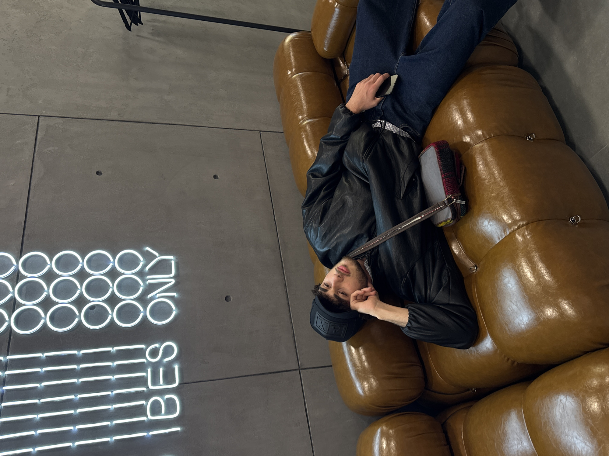 A man is lying on a brown leather sofa in Yerevan, Armenia. The walls are gray and a neon sign says "Vibes Only". The man is wearing a black leather jacket and jeans. He is holding a phone in his left hand and his right hand is resting on his head. He is wearing a black hat. He has a black shoulder bag. The sofa is segmented into large round cushions.