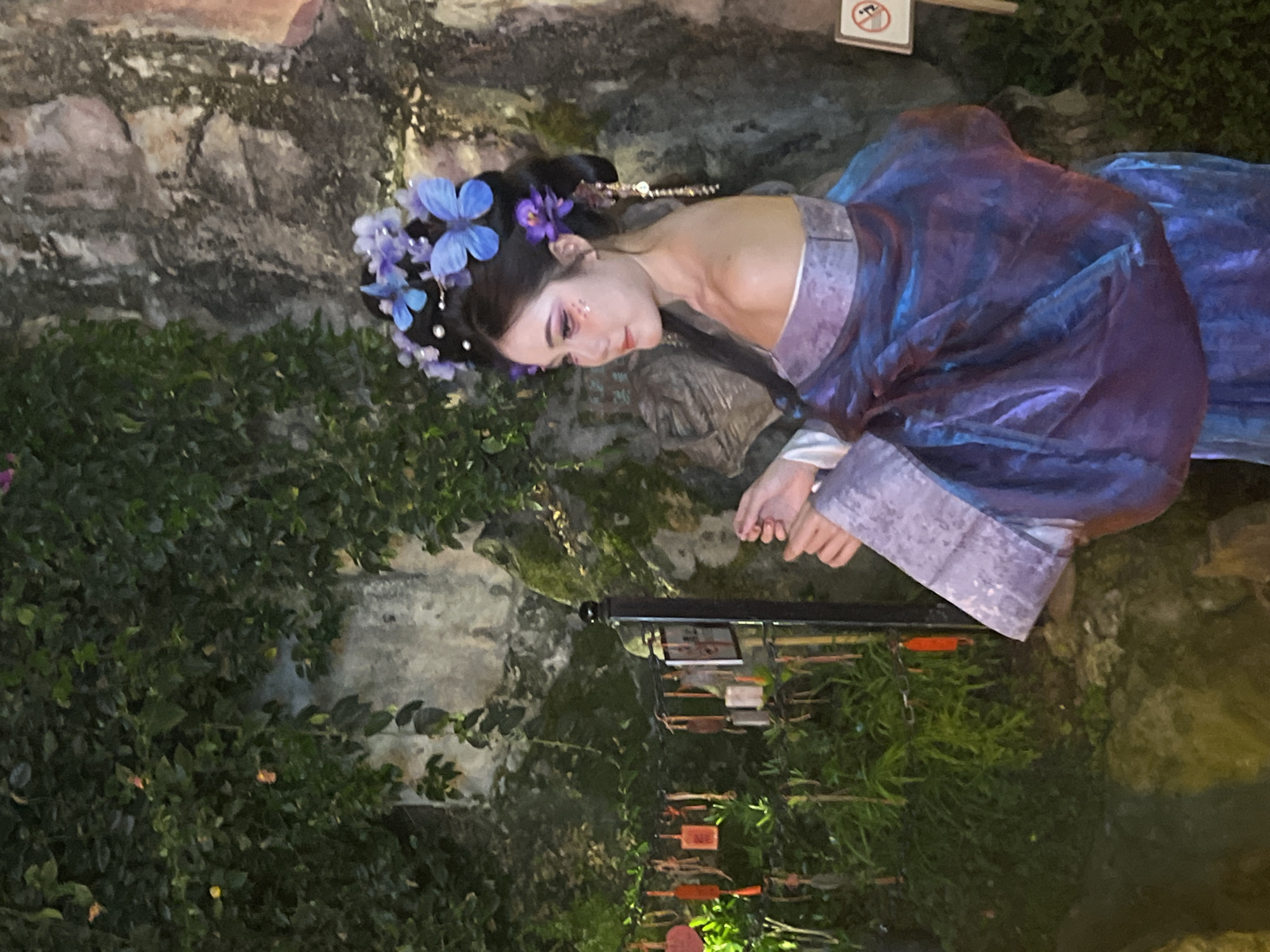 A woman in a traditional Chinese dress lies on a rock surface in a garden setting. She wears a dark wig adorned with blue and purple flowers and has delicate makeup on her face. Her dress is predominantly blue and purple with a silvery trim around the edges. She rests her hands near a small, dark structure with hanging objects, and the scene is surrounded by greenery. The image may have been taken at the Yuzhong District of China.