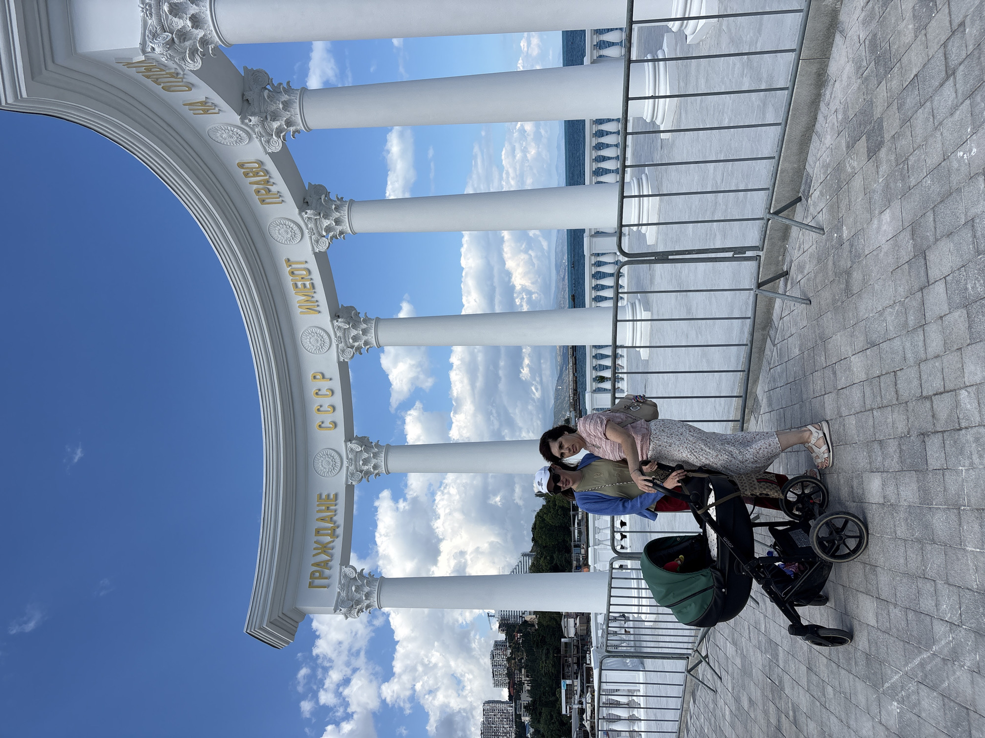 The image captures two women and a baby stroller positioned in front of an ornate white colonnade with gold lettering. The lettering on the structure reads "ГРАЖДАНЕ СССР ИМЕЮТ ПРАВО НА ОТДЫХ", which translates to "Citizens of the USSR have the right to rest." Behind the colonnade, the Black Sea is visible, with a cloudy blue sky overhead. In the background, a city with buildings is nestled on a hillside. The two women are standing close together, with one woman wearing a blue cardigan and the other a pink top. A green and black stroller is situated to the left of the women. The scene appears to be a public promenade or walkway in Alushta, Ukraine, likely a tourist or historical site.