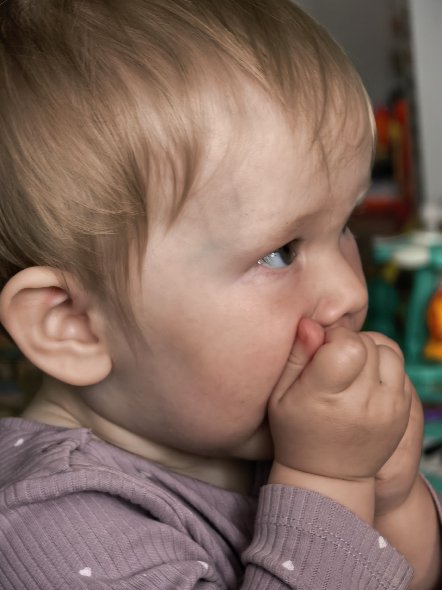The image shows a close-up, side profile of a young child, likely a toddler, with fair hair. The child is wearing a mauve, ribbed long-sleeved shirt with small white heart patterns. Their hands are clasped together near their mouth, with fingers tucked inside. The background is blurred, but appears to be an indoor setting with colorful toys visible on the right side. There is no indication of the location within Sterlitamak, Russia.