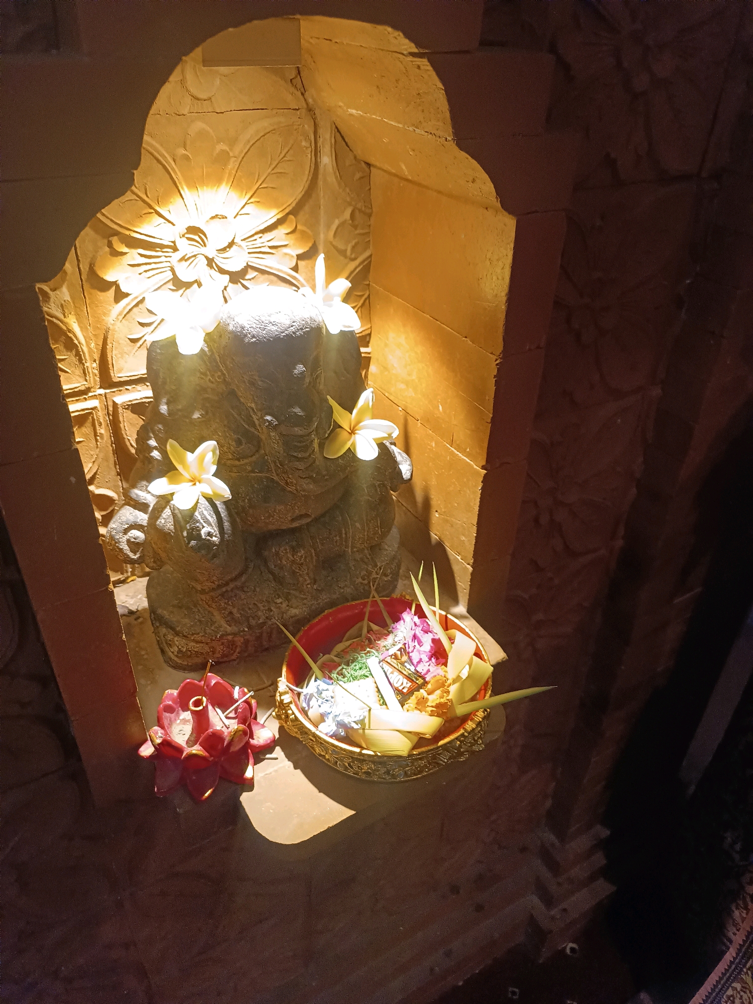 This image depicts a religious shrine, possibly dedicated to Lord Ganesha, located in Tabanan, Indonesia. The shrine is constructed of carved stone, with intricate floral patterns adorning the back wall. A stone statue of Ganesha sits at the center, decorated with several white and yellow plumeria flowers. In front of the statue is a woven offering basket filled with flowers and what appears to be incense sticks. To the left of the offering basket is a red, lotus-shaped incense burner, also containing incense. The lighting is warm and atmospheric, with a spotlight illuminating the Ganesha statue and the carved wall. The surrounding environment appears to be dimly lit, suggesting it might be an indoor or evening setting. The overall impression is one of a serene and spiritual space.