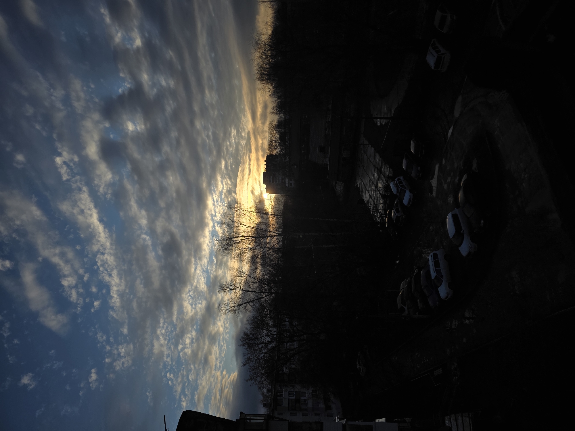 The image shows a view of an urban environment at dusk or dawn. The sky is filled with clouds, illuminated by a golden light source, likely the sun. Below the sky, the image shows a silhouette of trees and buildings. On the right side of the image, a parking lot with several cars is visible, though largely obscured by shadow. The location appears to be a city, but specific details are not discernible. There are no people visible in the image.