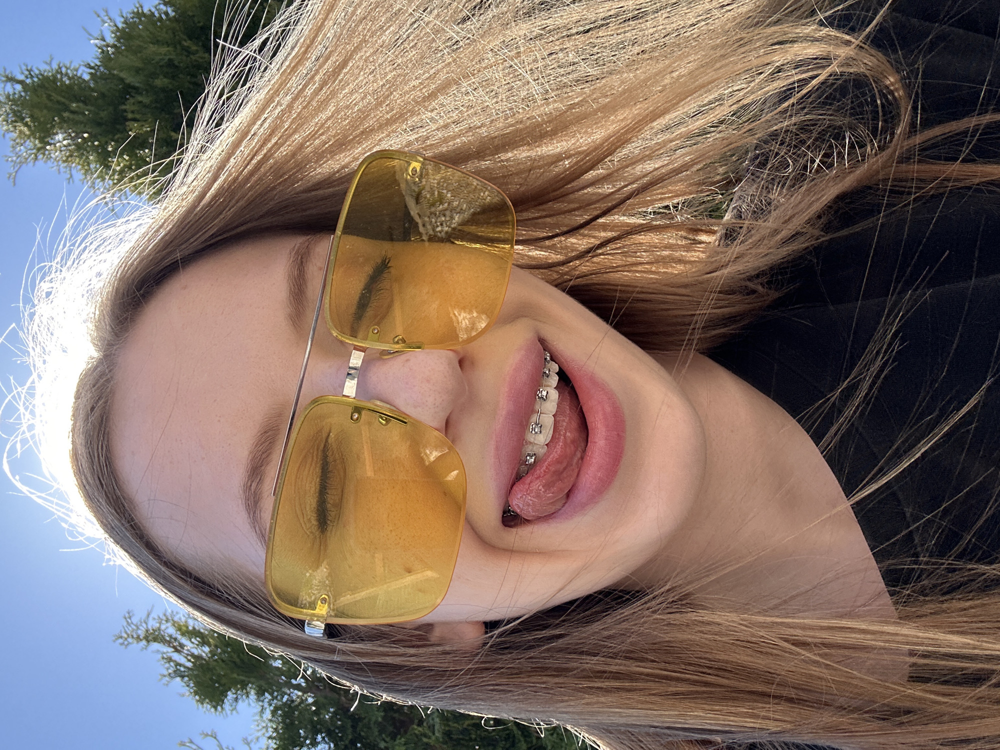 A young woman with long blonde hair and wearing yellow sunglasses poses in what appears to be an outdoor setting. She is wearing braces on her teeth and sticking her tongue out. The environment includes a glimpse of green foliage and a bright blue sky, suggesting a sunny day. The photo could have been taken in the city of Chaikovsky, Russia.