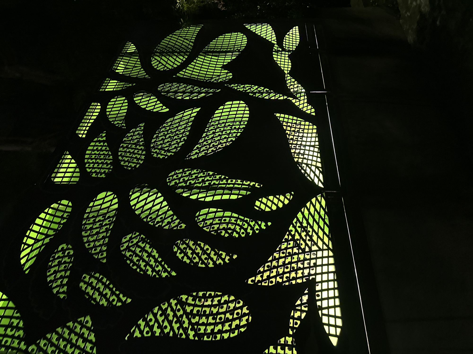 The image shows a close-up of a decorative metal screen illuminated from behind with green light. The screen is designed with cutouts in the shape of leaves, some of which have a criss-cross pattern within them, resembling veins. The background is dark, suggesting nighttime. The screen is mounted on a dark, solid structure that appears to be a wall or fence, with a thin horizontal railing visible in front of it. The overall impression is of an artistic outdoor installation, possibly in a park or along a walkway. Without additional context, the specific location within Singapore, Singapore, cannot be determined.