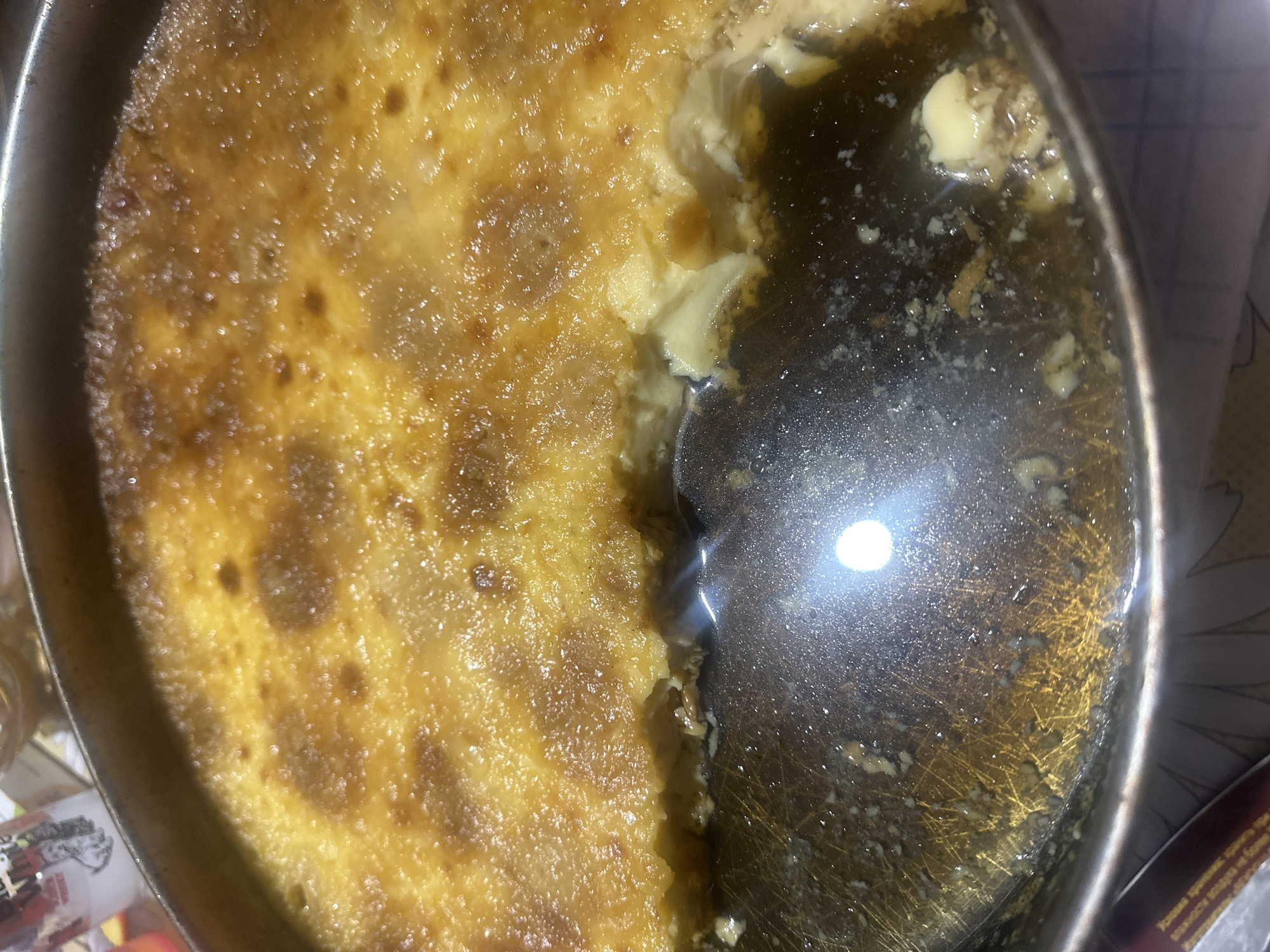 A slightly aerial view captures a partially consumed baked custard, possibly crème caramel or flan, in a round metal baking pan. A portion has been scooped out, revealing a syrupy liquid at the bottom. The custard has a browned, slightly uneven surface. The pan sits on a surface, part of which is visible and out of focus. There is no indication of people or a specific location like Razhevo Konare, Bulgaria. The scene depicts a dessert that has been eaten from, in a pan on a surface.
