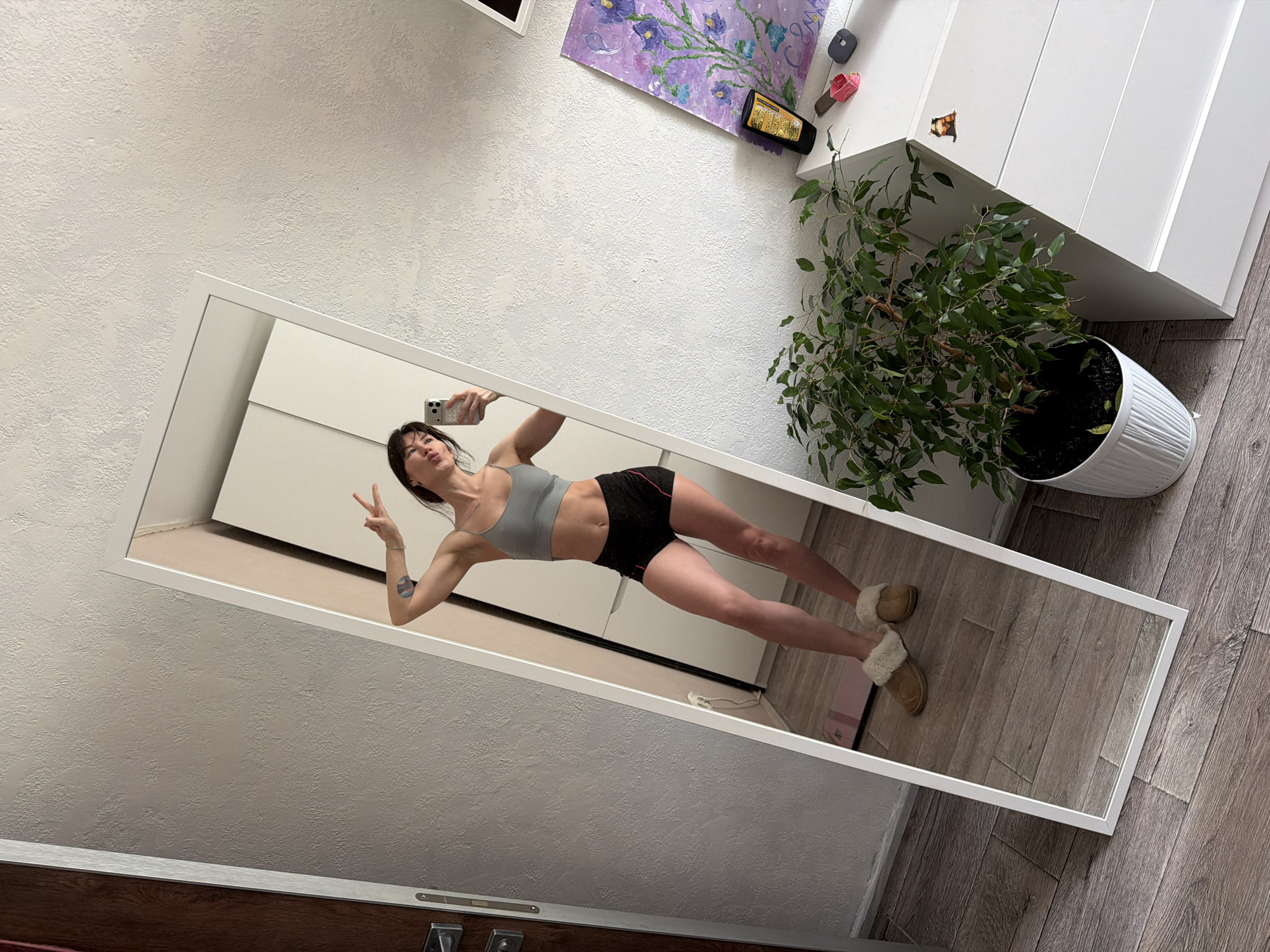 A woman is taking a selfie in a full-length mirror in what appears to be a home in Perm, Russia. The woman is wearing a grey sports bra and black shorts, and she is flashing a peace sign with one hand while holding a smartphone in the other. Her hair is dark and pulled back. The reflection shows her standing on a wooden floor with a pink yoga mat nearby. In the background, there's a white dresser with a houseplant on top, as well as a painting on the wall. The apartment has white walls. The mirror is leaning against the wall.
