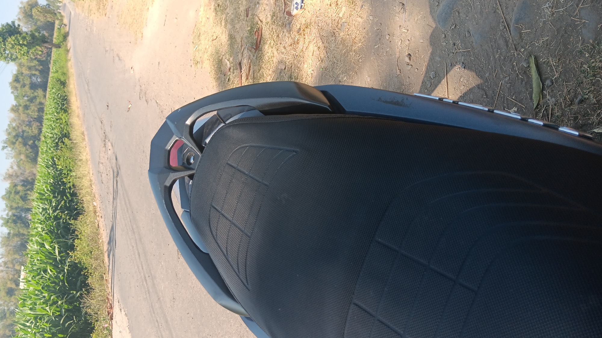 The image shows the seat of a motorcycle parked on the side of a rural road in Gemenggeng, Indonesia. The road is paved and appears to be in fair condition, with some cracks and potholes. To the left of the road is a field of corn, lush and green, stretching towards a line of trees in the background. The sky is clear and blue. The motorcycle seat is black with a textured pattern. The overall scene depicts a quiet, sun-drenched rural landscape.