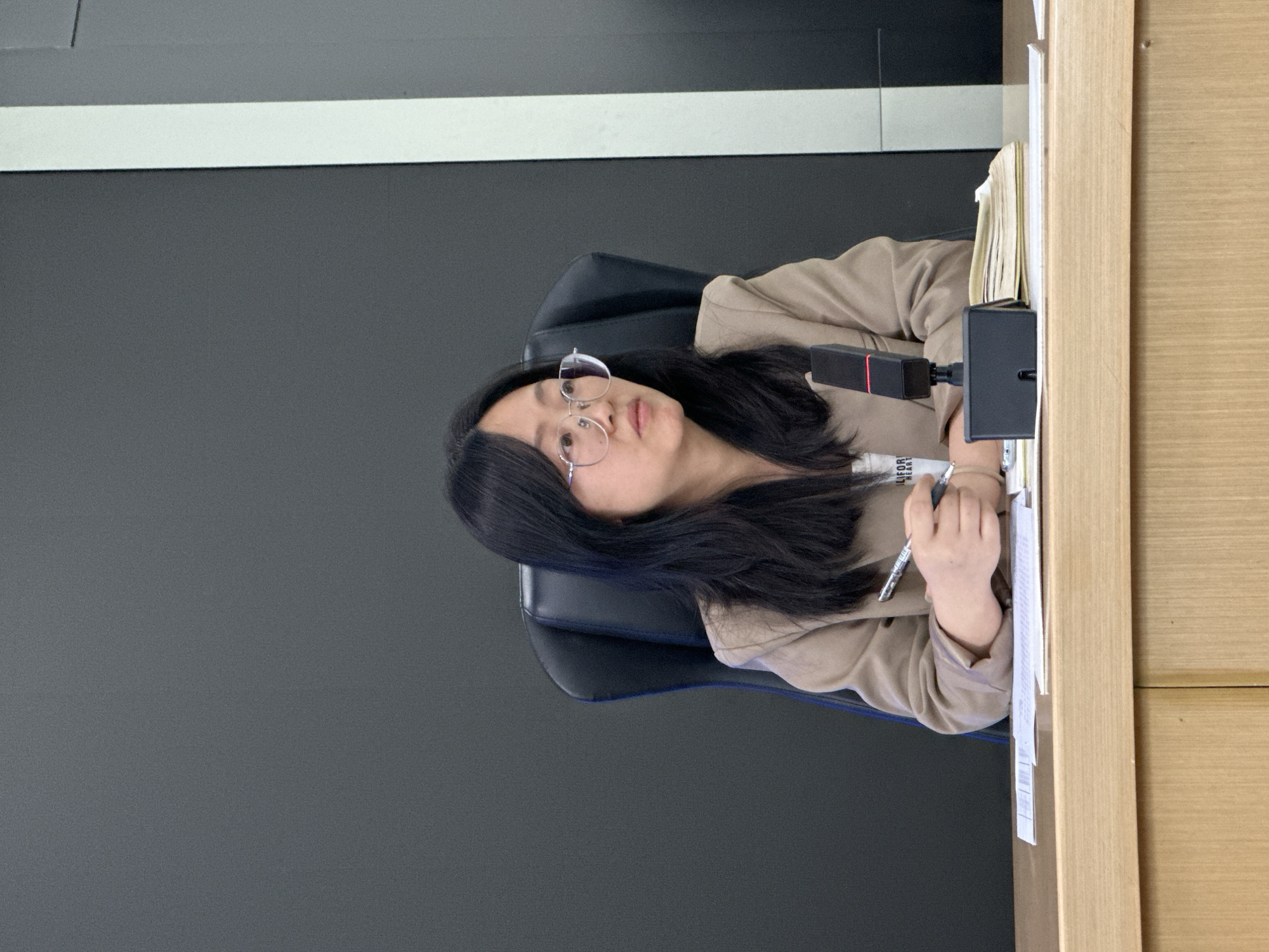 A woman is seated in a blue office chair at a desk. She is wearing glasses and a beige blazer. She has long dark hair. On the desk is a microphone, a book, and some papers. She is holding a pen in her hand and appears to be writing or taking notes. The background consists of a gray wall with a white horizontal stripe near the top and a light wood panel on the right side. There is no information in the image to confirm or deny the location in the city of Jiangjin, China.