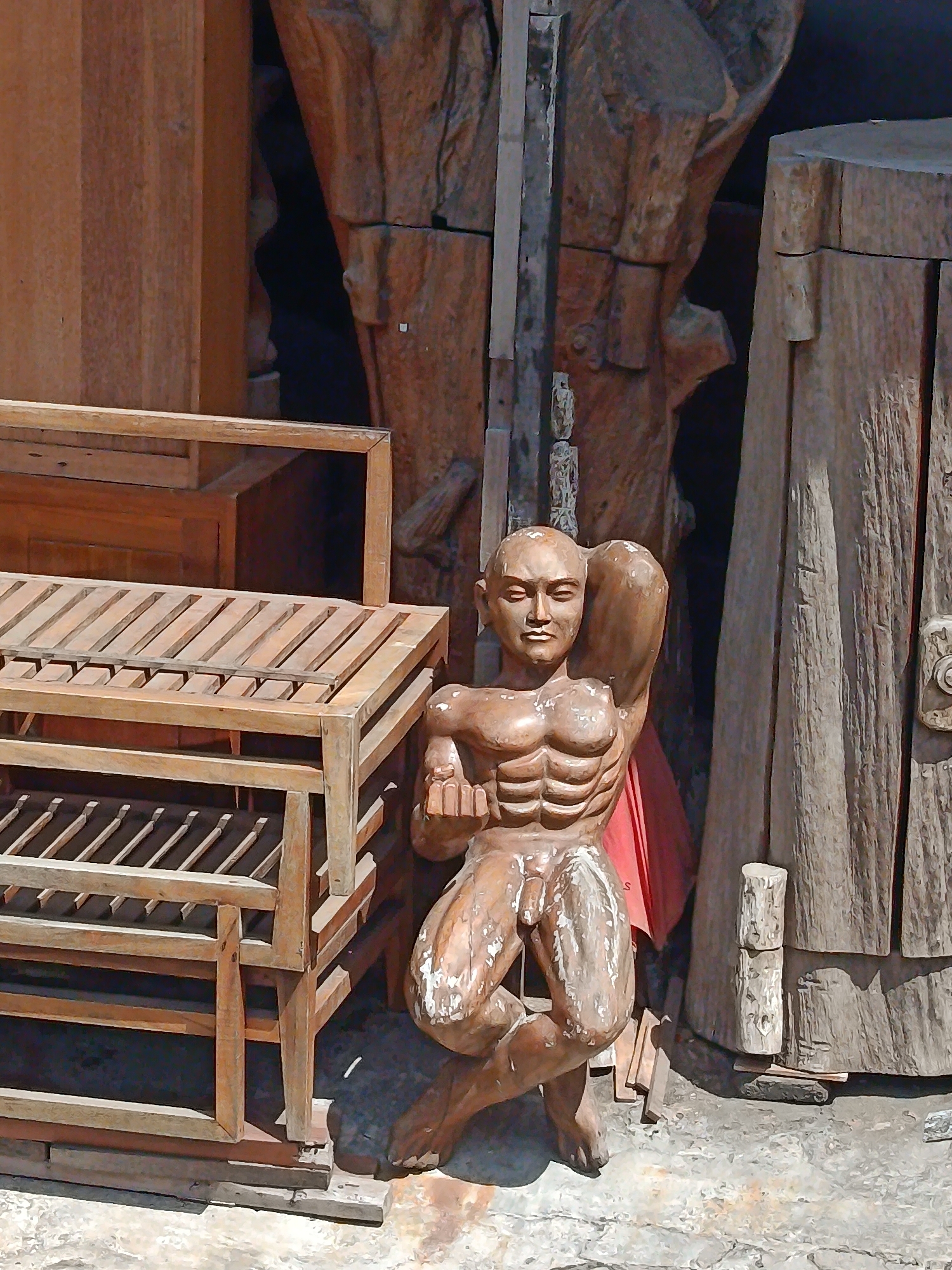 The image depicts a carved wooden statue of a muscular, nude man with his left arm raised behind his head and his right arm bent at the elbow with his fist held near his chest. The statue is positioned in front of large, weathered wooden structures and a slatted wooden display rack. The statue has a detailed carving of abdominal muscles and a visible phallus. The wood of the statue appears to be aged and has some white paint or residue on its surface. The environment suggests an outdoor setting, possibly a market or artisan's stall, with the ground appearing to be concrete or paved. This statue is located in Kuta, Indonesia, an area known for its vibrant arts and crafts scene.