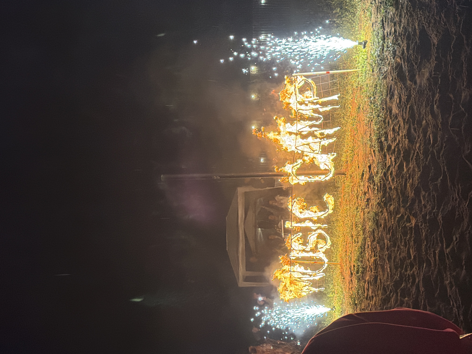 The image depicts an outdoor nighttime scene, likely at an event. In the foreground, a person wearing a maroon garment is partially visible on the left. The main subject is a large sign spelling out "MUSIC CAMP" that is engulfed in flames. The sign is constructed of metal letters and is supported by a metal frame. On either side of the flaming sign, there are pyrotechnic fountains, one on the left and another on the right, emitting sparks and light. In the background, there is a white tent with people gathered underneath it, and further back, there are more lights that might indicate buildings or other structures. The ground appears to be a mix of sand and grass. The sky is dark and appears to have some faint vertical streaks, possibly indicating rain or falling debris from the pyrotechnics. While the image does not contain specific visual cues to pinpoint the location to Menzelinsk, Russia, it captures a festive and dramatic event.
