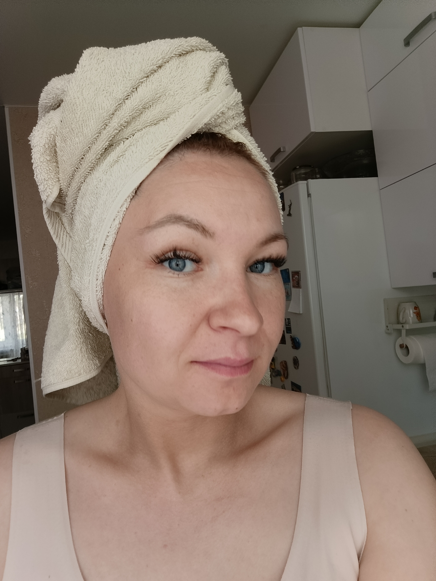 A woman with a white towel wrapped on her head is standing indoors, likely in a kitchen. She has blue eyes with prominent eyelashes. She is wearing a light beige tank top. Behind her are white kitchen cabinets and a refrigerator with magnets attached. The location is possibly in Chaikovsky, Russia, based on prior knowledge.