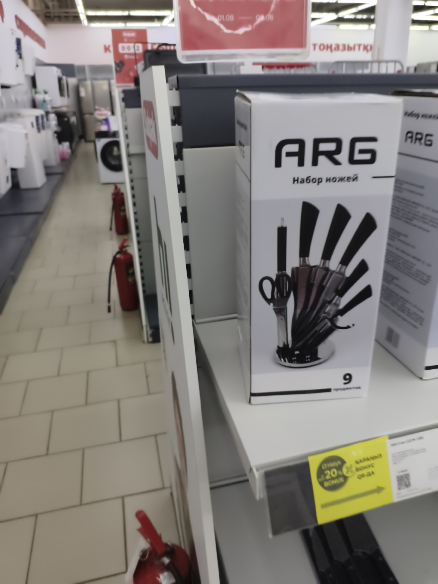 This is a medium-angle, eye-level, slightly blurred, indoor shot of a retail store aisle in Aktobe, Kazakhstan. The aisle is lined with shelves displaying various household appliances and kitchenware. In the foreground, a white cardboard box with the brand name "ARG" printed in large black letters and the Russian text "Набор ножей" (Knife Set) is prominently displayed. The box features a graphic illustration of a knife block with several knives and a pair of scissors. Below the illustration, the number "9" is visible, likely indicating the number of items in the set. To the left of this box, another identical box is partially visible. On a shelf below the knife set boxes, a yellow promotional sticker with Russian text and a QR code is attached. Further down the aisle, two red fire extinguishers are visible, one closer to the foreground and another further back. The flooring appears to be tiled. In the background, more shelves with appliances, including what look like washing machines and refrigerators, can be seen. The overall impression is of a well-stocked appliance and home goods store.