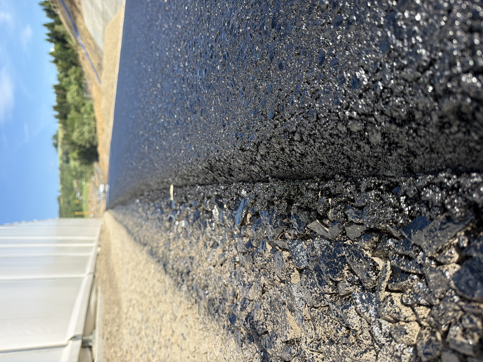 The image is a low-angle, close-up shot of a newly paved asphalt surface bordering a construction site. On the left, a rough, unpaved area with gravel and dirt is visible, adjacent to a white, tent-like structure. The dividing line between the unpaved area and the fresh asphalt is sharp. The asphalt surface is dark, wet, and reflects the sunlight, creating numerous small, bright highlights. In the background, a hill covered with trees and a clear blue sky can be seen. There are no people or vehicles prominently featured in the immediate foreground. The scene appears to be a section of road construction in an outdoor environment, possibly in a rural or suburban area, given the natural landscape in the background. The location is identified as Janowice, Poland.