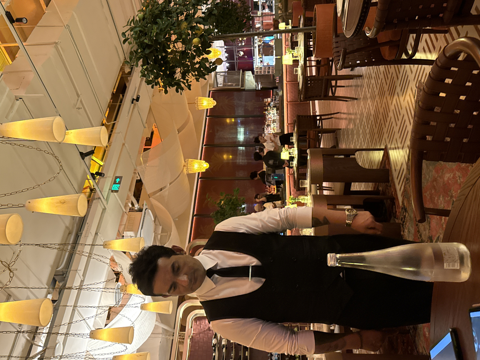 A server stands in a restaurant, looking down at a bottle of water on a table. He wears a white dress shirt, a black vest, and a black tie. The restaurant has a modern interior design with hanging pendant lights, a bar in the background, and several tables and chairs. The floor has a geometric tile pattern. The image appears to be taken in Yiwu, China, as indicated by the presence of what looks like a restaurant within a commercial complex commonly found in Yiwu.