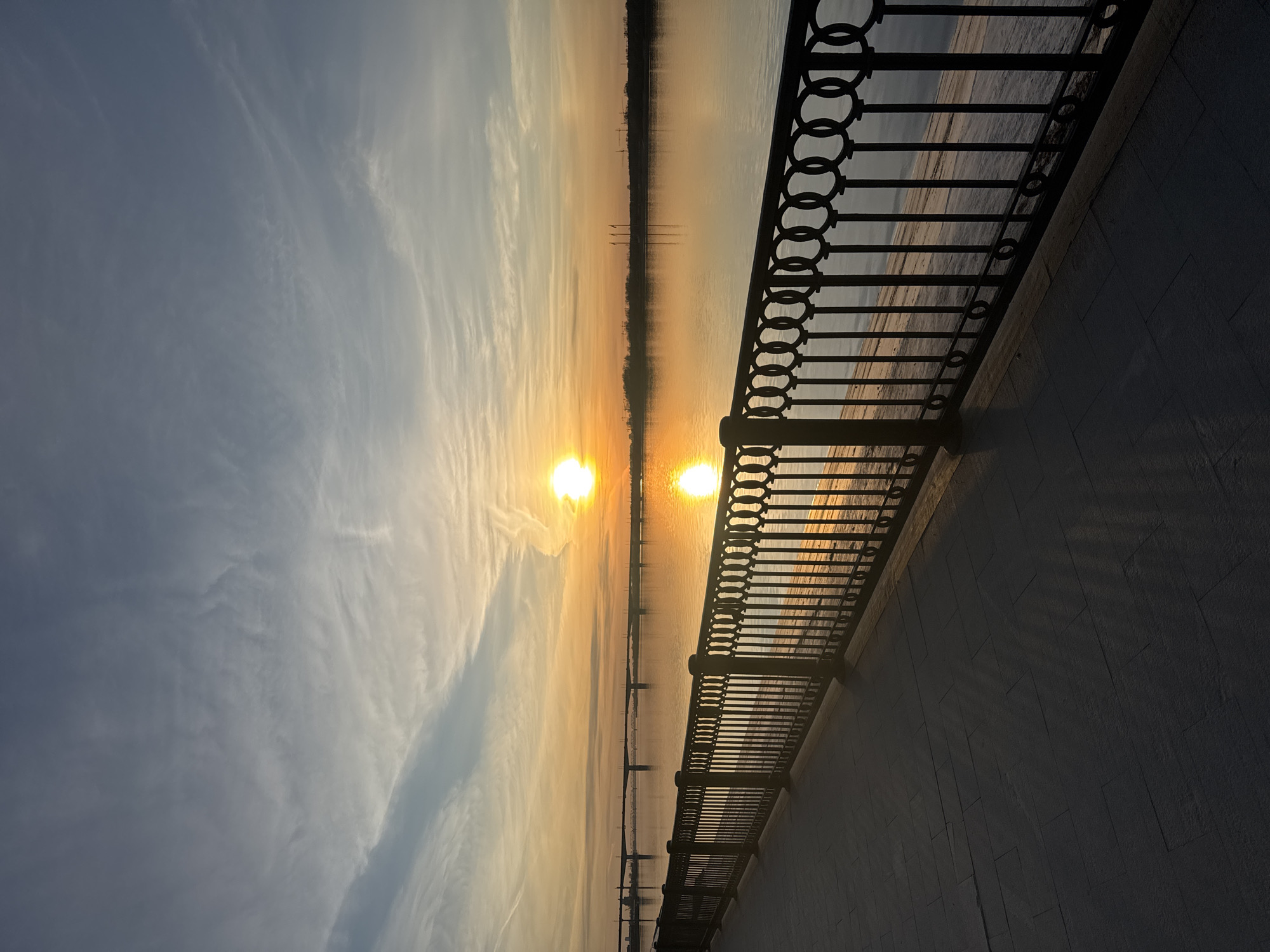 The image displays a serene landscape at either sunrise or sunset, captured from an embankment overlooking a wide body of water. The photograph is oriented horizontally, showcasing a vibrant sky, calm water, and a distinctive railing.
A bright, glowing sun is prominently positioned in the sky towards the center-right, casting intense orange and yellow light across the horizon. Its equally radiant reflection stretches as a bright path across the tranquil water below. The sky exhibits a gradient, transitioning from a soft blue-grey on the far left to warm golden and orange hues near the sun, with delicate, wispy clouds scattered throughout.
Along the right side of the frame, a dark, ornate metal railing with repeating circular motifs runs parallel to the water's edge, separating it from a paved promenade or embankment. The railing is silhouetted, and its shadow, along with the shadows of its vertical posts, extends diagonally across the textured, grey-tiled ground of the promenade, indicating a low sun angle. The embankment itself is largely in shadow.
On the distant horizon across the water, a faint, dark line suggests the opposite bank, with subtle, indistinct features that could be trees or buildings. The overall scene conveys a sense of peaceful natural beauty, with no human activity visible. No text is discernible in the image. Given the location context, this is likely an embankment along the Kama River in Perm, Russia.