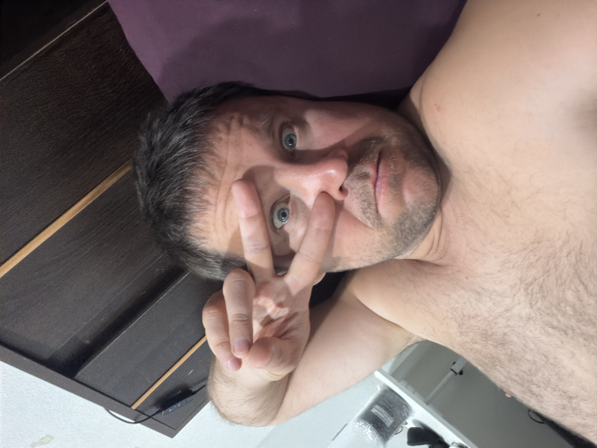 A man is lying in bed and taking a selfie. He is looking at the camera with his eyes open and his hand in a peace sign in front of his face. The man has short, dark hair and a beard. He is shirtless and his chest is covered in hair. The background is a dark wooden headboard of a bed and a purple pillow. To the left of the man, there is a white shelf with a black container on it. The image appears to be taken in Baan Mae Nam, Thailand.