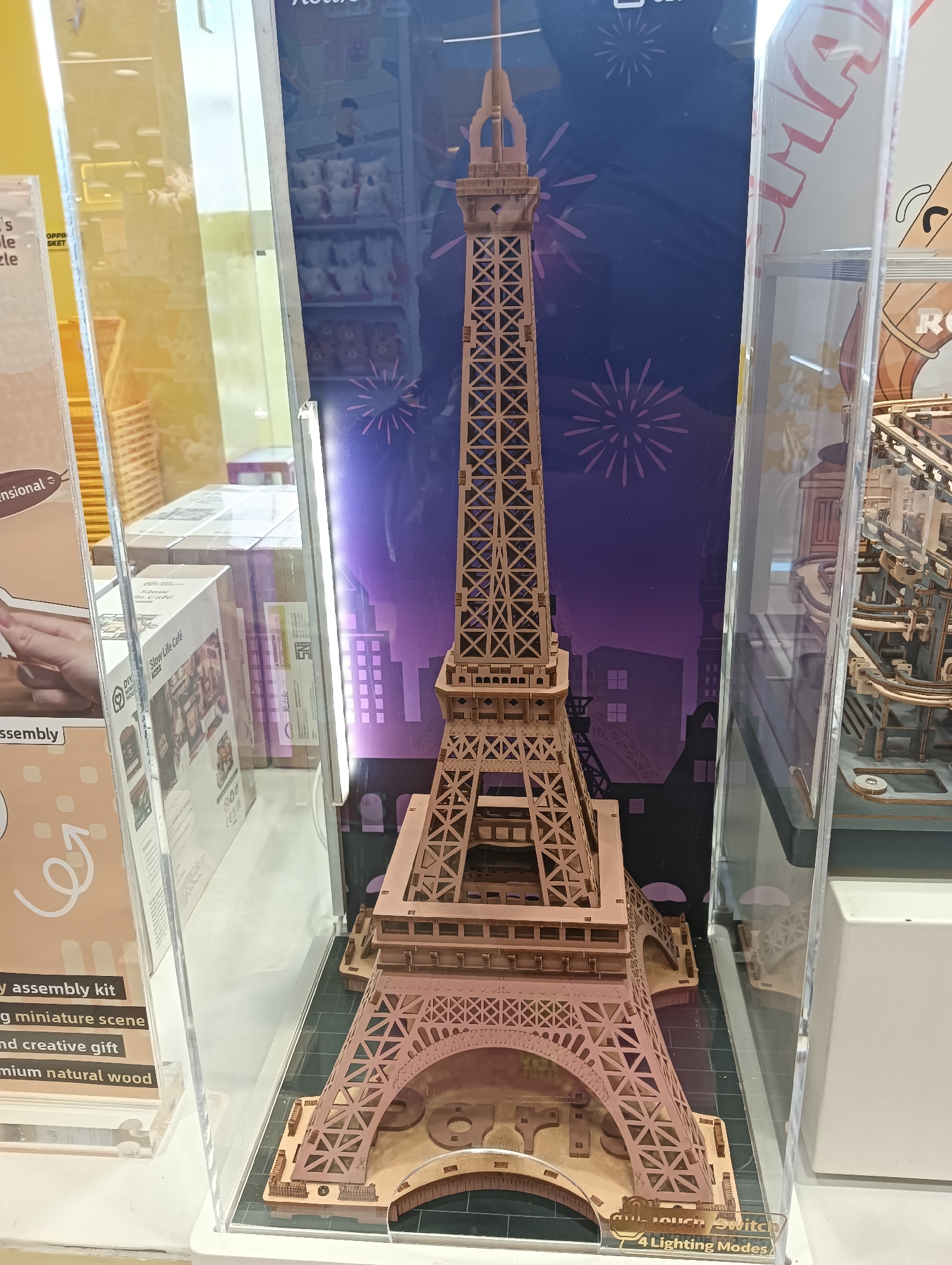 The image shows a detailed wooden model of the Eiffel Tower displayed in a clear acrylic case. The model is intricately designed with latticed structures and appears to be a DIY assembly kit. Behind the Eiffel Tower, there's a backdrop featuring a cityscape with silhouetted buildings and purple and pink fireworks. To the left of the Eiffel Tower, part of a display with text describing an "assembly kit" and "creative gift" made from "premium natural wood" is visible. To the right, another display case contains a wooden roller coaster model. The overall setting appears to be a retail store or exhibition. The location is specified as Kediri City, Indonesia.
