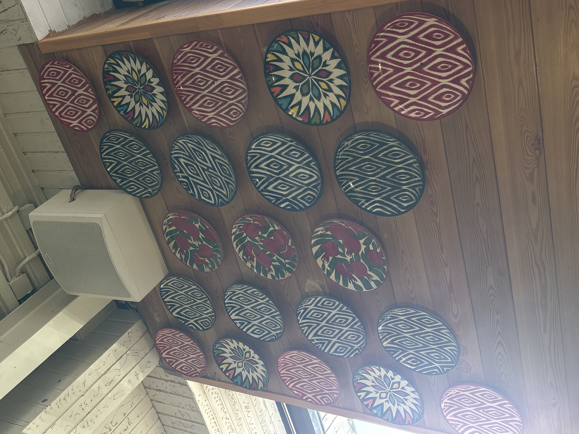An interior shot shows a wooden wall adorned with numerous oval, decorative plates. The plates feature a variety of patterns, including geometric designs in red and white, blue and white, and colorful floral or starburst motifs. A white speaker is mounted on the wall above the plates. The ceiling, made of wooden beams and planks, is visible at the top of the image. The scene appears to be inside a building, possibly a restaurant or home, in the city of Solnechnoye, Russia, judging by the style of the interior elements and the context provided.