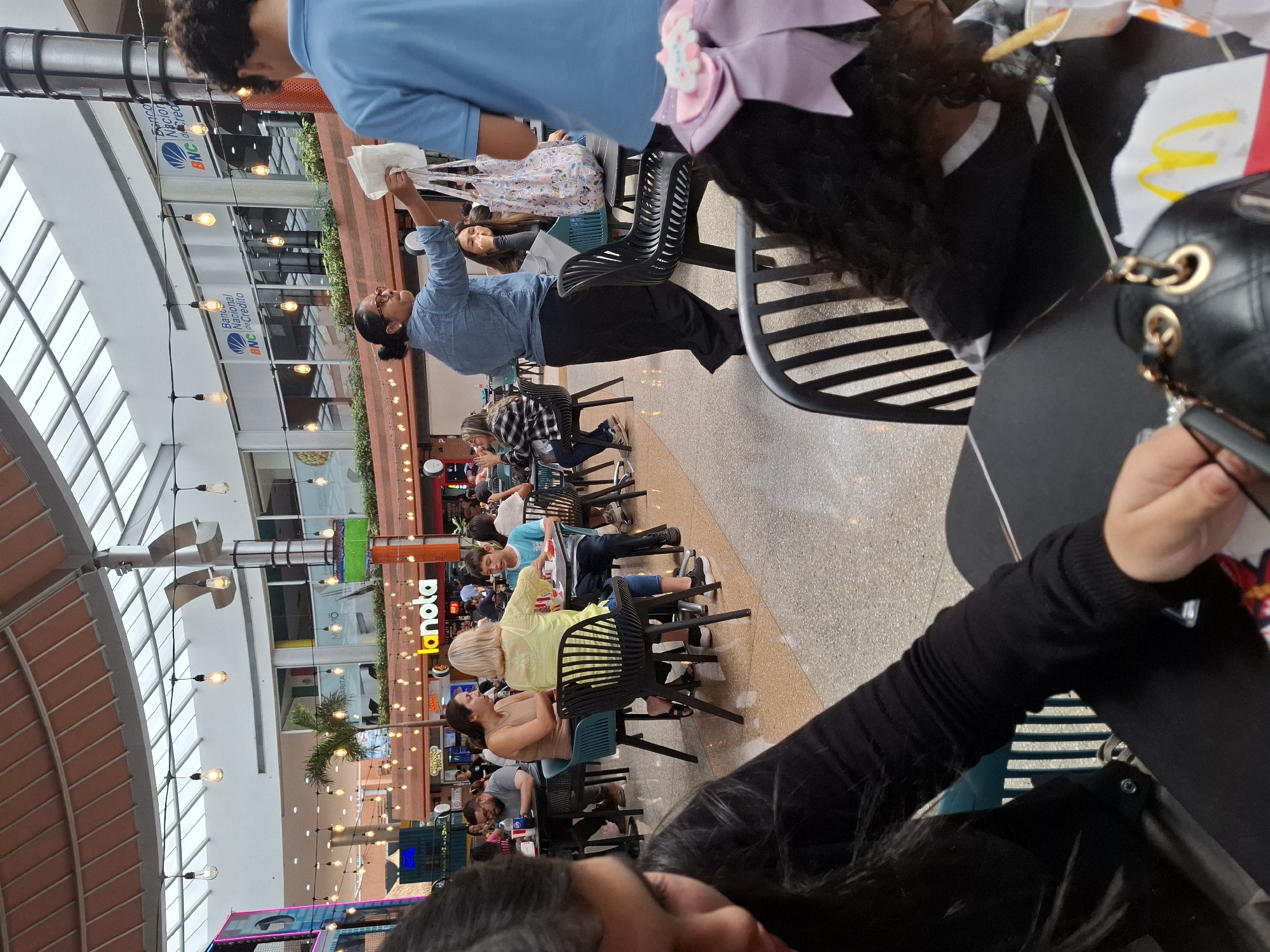 The image depicts a busy food court inside a shopping mall, likely in Pampatar, Venezuela, given the presence of "La Nota" signage, a known establishment in that region. In the foreground, a young girl with long dark hair and a black top is partially visible, her hand reaching out towards the table. To her right, another child with dark curly hair and a large light purple bow is seated at a table, with a cup of french fries and a McDonald's bag visible.
Further into the scene, a woman wearing a blue denim shirt and black pants is holding a white bag and appears to be interacting with someone. Several other individuals are seated at tables, eating and conversing, including a woman in a light yellow top and a boy in a blue shirt. The food court is illuminated by string lights and natural light filtering through a large skylight. The overall environment suggests a public, casual dining space within a commercial establishment.