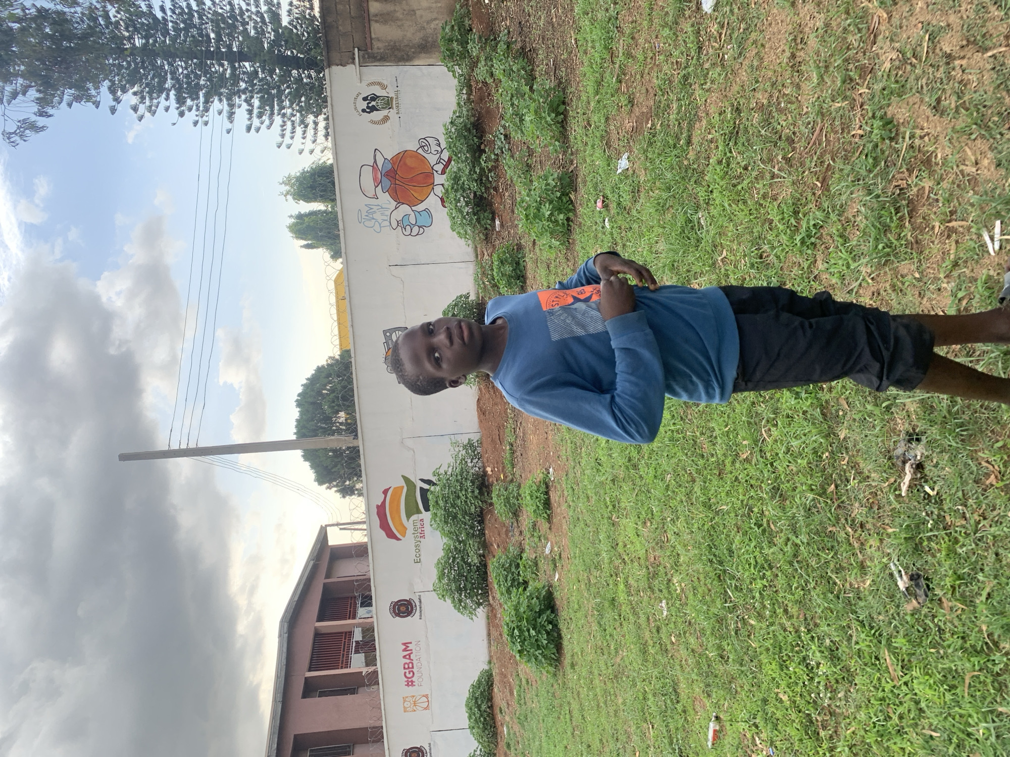 A young male stands on a grassy area next to a painted wall, seemingly in Gura Top, Nigeria. The wall features cartoon basketball imagery and logos, including "Ecosystem Africa" and "#GBAM FOUNDATION." The boy wears a blue long-sleeved shirt and dark shorts. Background details include a building, power lines, a tree, and a cloudy sky. There's litter scattered on the grass.