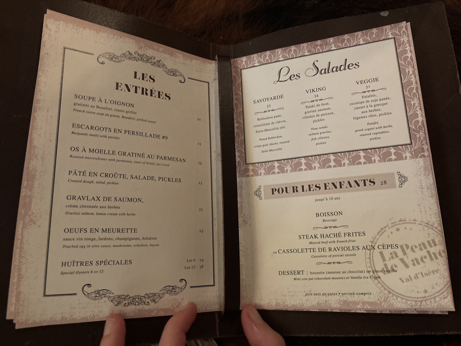 The image displays an open menu, held by a person's hands, in what appears to be a dimly lit restaurant setting. The background is dark and out of focus. The menu has a dark brown cover and two light-colored pages with decorative borders.
The left page is titled "LES ENTRÉES" (Starters) and lists several French dishes with their prices in euros. Items include "SOUPE À L'OIGNON" (French onion soup), "ESCARGOTS EN PERSILLADE #9" (Burgundy snails), "OS À MOELLE GRATINÉ AU PARMESAN" (Roasted marrowbones), "PÂTÉ EN CROÛTE, SALADE, PICKLES", "GRAVLAX DE SAUMON", "OEUFS EN MEURETTE" (Poached egg in wine sauce), and "HUÎTRES SPÉCIALES" (Special Oysters). Prices range from 19 to 38 euros.
The right page is divided into two main sections: "Les Salades" and "POUR LES ENFANTS" (For Children). The salads offered are "SAVOYARDE" (with Reblochon and goat cheese), "VIKING" (with tuna tataki, salmon gravlax), and "VEGGIE" (with falafel and roasted vegetables), priced at 33, 34, and 31 euros respectively. The children's menu, for those up to 10 years old and priced at 28 euros, includes a beverage, a choice of "STEAK HACHÉ FRITES" (Minced beef with French fries) or "CASSOLETTE DE RAVIOLES AUX CÈPES" (Cassolette of porcini ravioli), and a dessert of "bouette (mousse au chocolat)" or vanilla ice cream.
A watermark on the right page reads "La Peau de Vache Val d'Isère," indicating the restaurant's name and its location in Val-d'Isère, France. The bottom of the menu states "prix nets, en euros * service compris" (net prices, in euros * service included).