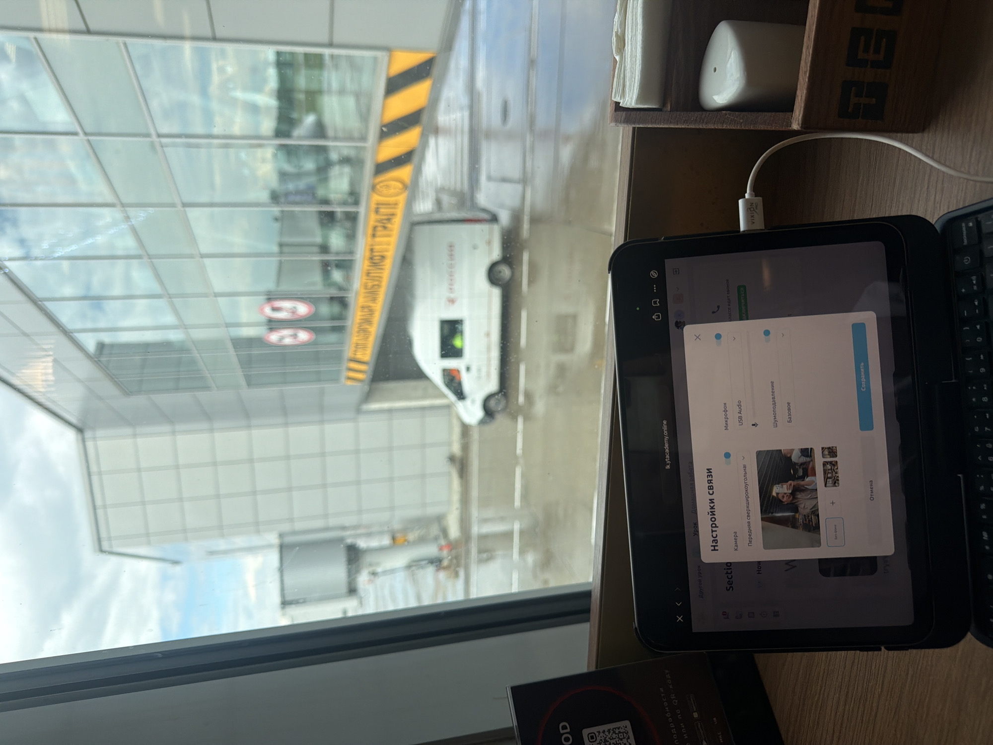 An interior view from an airport waiting area shows a tablet computer displaying a video conferencing interface, with the tablet placed on a keyboard. Outside the window, an airport jet bridge and a white service van are visible, with the ground appearing wet. In the foreground, a wooden napkin holder and a salt shaker are present. The tablet displays "Настройки связи" (Connection Settings) with options for Camera, Microphone, and Sound. The webcam preview shows a woman taking a selfie. The overall setting suggests a traveler using technology while waiting for a flight.
