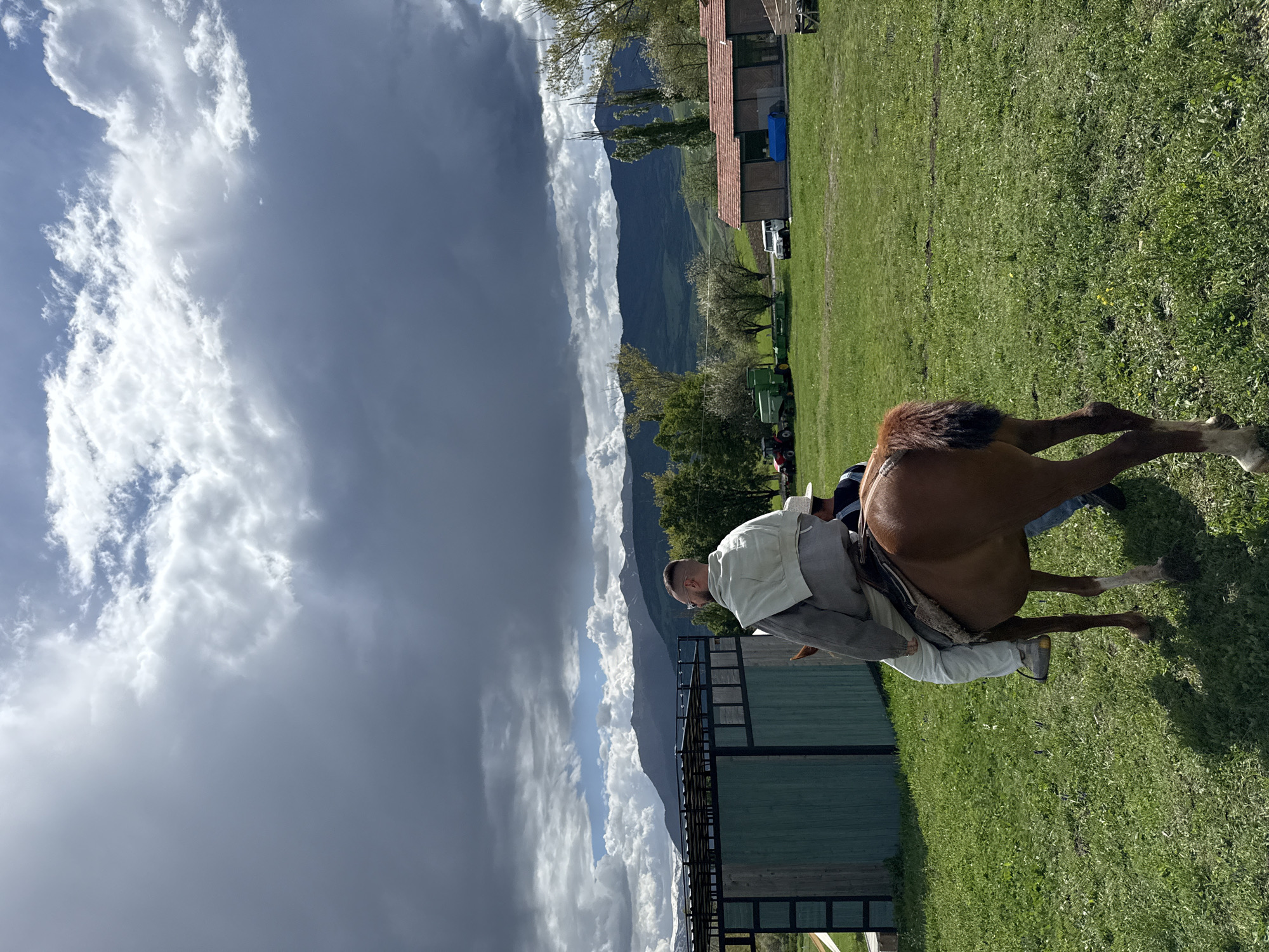 The image shows a man on horseback in a grassy field. He is wearing light-colored clothing and the horse is brown. In the background, there are mountains, clouds, and some buildings. The location is Dsegh, Armenia. There is a fence in the middle ground. A dark storm cloud is prominent in the sky. A tractor is visible to the rear. The horse is standing still.