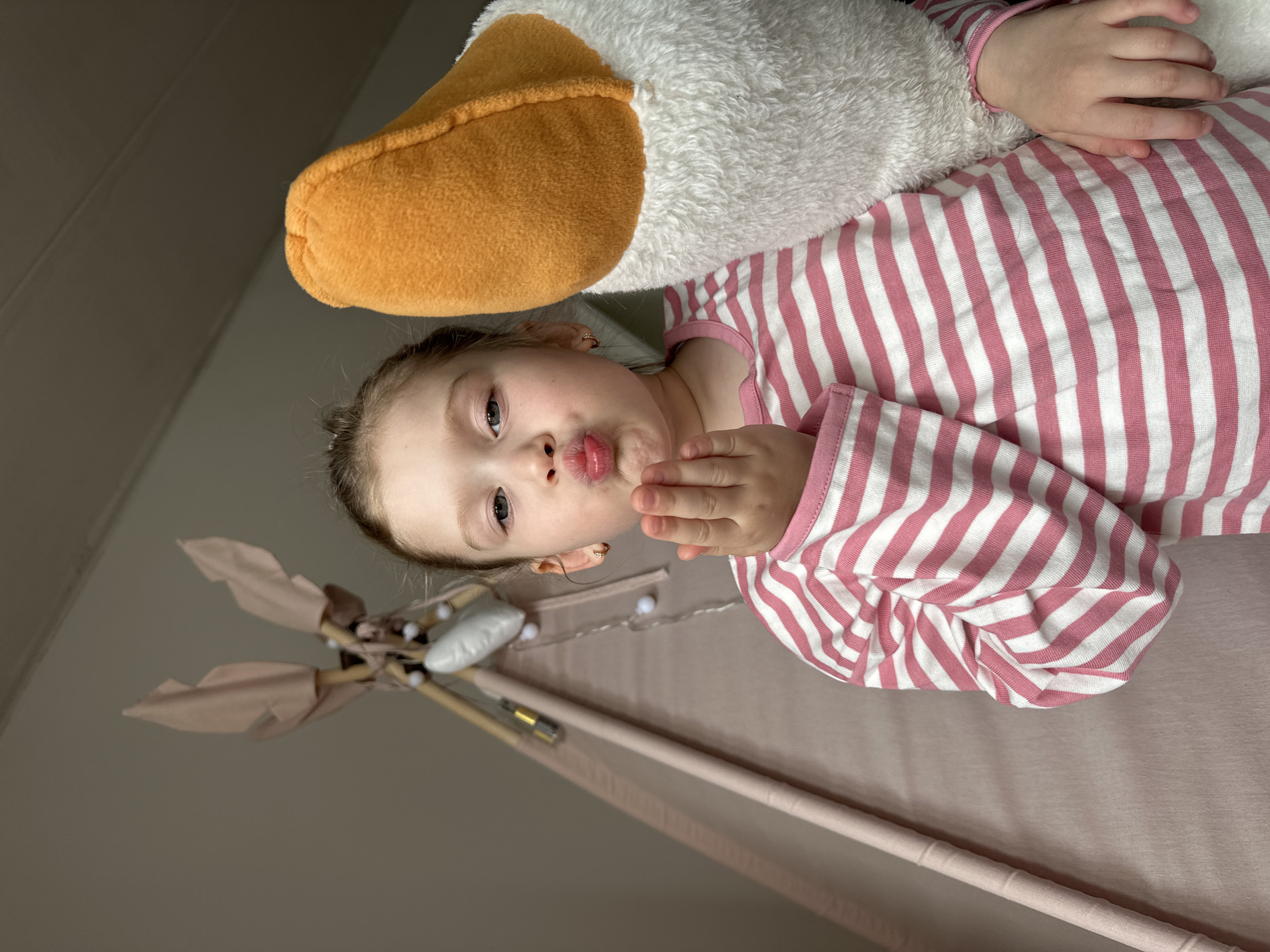 A young child lies in a pink teepee, clutching a plush white goose with an orange beak. The child wears pink and white striped pajamas and puckers their lips, holding a hand near their face. The teepee is decorated with small white lights. The scene appears to be indoors, possibly a bedroom. There is no evidence to indicate that this image was taken in the city of Chaikovsky, Russia.