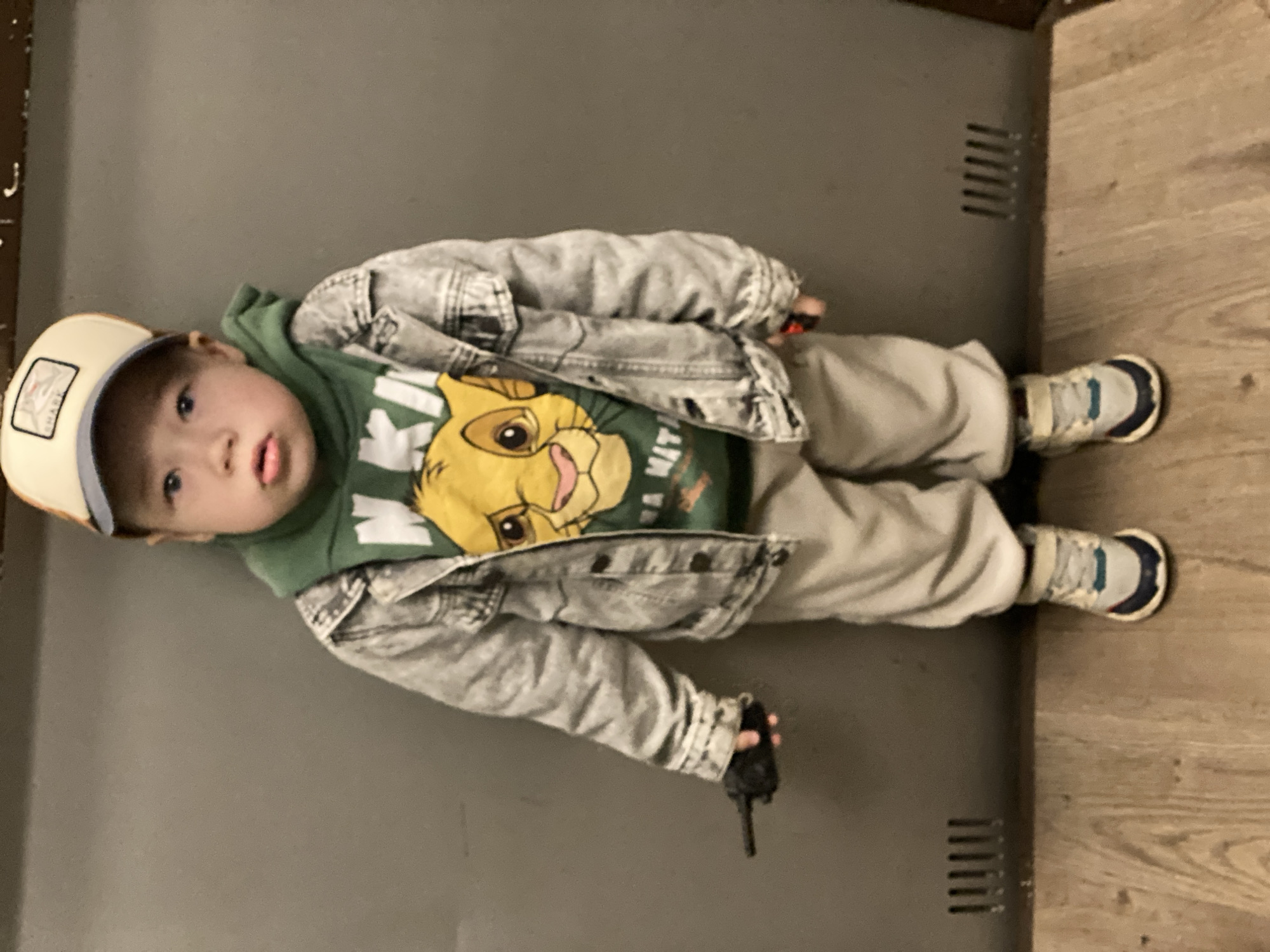 A young child, approximately 2 years old, is lying on a gray surface next to a wall with ventilation slots and an adjacent wooden floor. The child wears a cream-colored baseball cap, a green hooded sweatshirt with a "Lion King" graphic, a faded gray denim jacket, and beige pants. The child has light-colored sneakers with dark blue accents on their feet. The child is holding a black object resembling a walkie-talkie or microphone in their left hand. There is no indication of the location being Sochi, Russia.