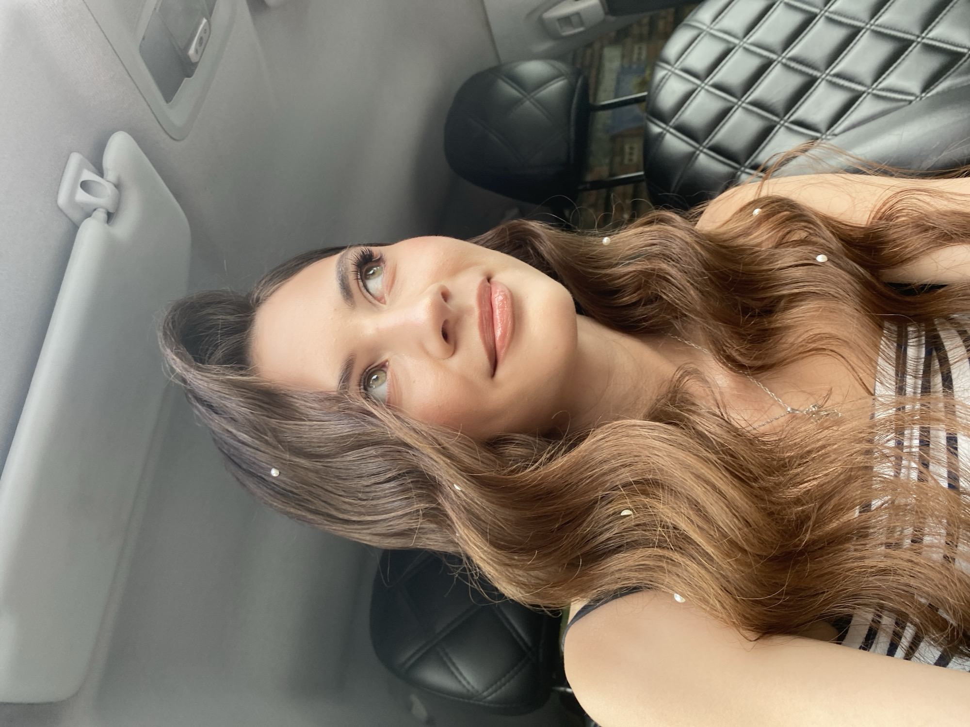 A young woman with long, wavy brown hair adorned with small white pearls is sitting in the passenger seat of a car. She is looking upwards and to her right with a slight smile. Her eyes are green, and she is wearing a striped top and a silver necklace. The car's interior is visible, including quilted black leather headrests and the car's ceiling with a sun visor. The scene appears to be captured during daylight. No specific location within Votkinsk, Russia is identifiable from the image.