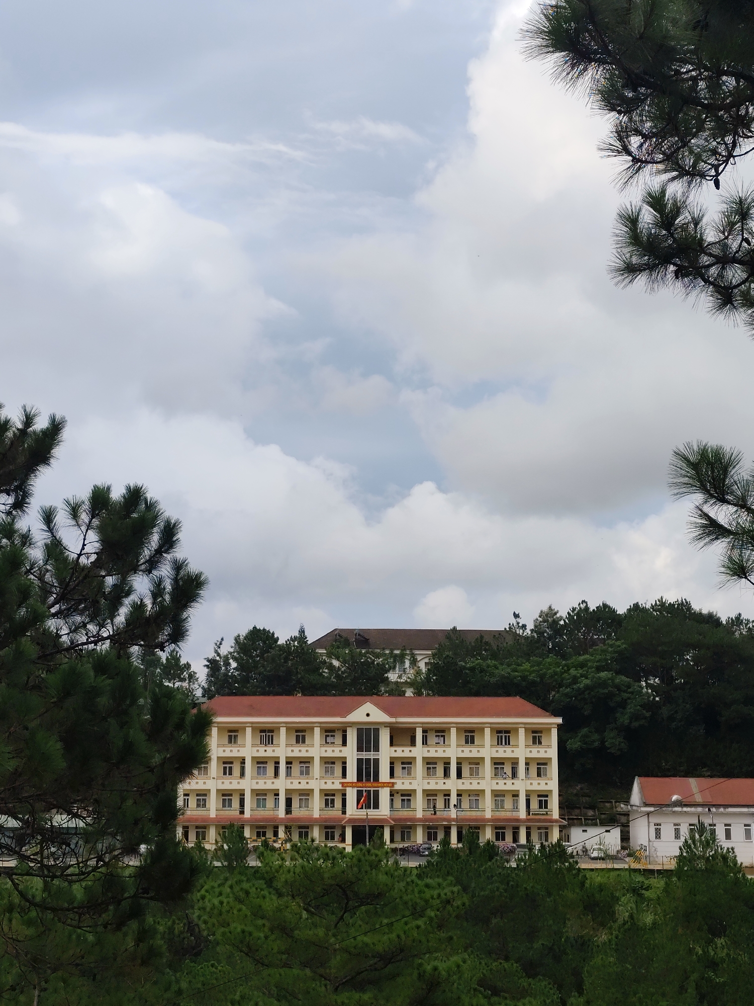 The image captures a serene, daytime scene in a hilly, forested landscape in Lạc Dương, Vietnam. A large, cream-colored building with a prominent red-tiled roof stands centrally, nestled amongst numerous dark green pine trees.
The main building is multi-storied, featuring at least five visible floors with multiple rectangular windows, some equipped with small balconies. Its architecture is functional, typical of an institutional building. In front of the building, a flagpole holds a red and yellow flag, likely the national flag of Vietnam. Partially visible parked vehicles suggest activity, though no people are explicitly seen.
To the right of the main building, a smaller, white building with a matching red roof is visible, situated further back. Another building, with a darker roof, can be glimpsed on a higher elevation behind the central structure, mostly obscured by the dense tree line. The surrounding environment is dominated by lush pine forests, with their characteristic long needles and some visible pinecones, giving the scene a fresh, natural feel.
The sky is overcast with large, diffused white and grey clouds, indicating a cloudy day with soft, indirect lighting. The overall atmosphere is tranquil and peaceful.
Visible text on a sign or banner on the front of the main building reads "TRƯỜNG TRUNG HỌC PHỔ THÔNG...", which translates to "High School...". This strongly suggests the building is a high school, possibly Lạc Dương High School given the provided location context. The remaining text is too blurry to decipher.