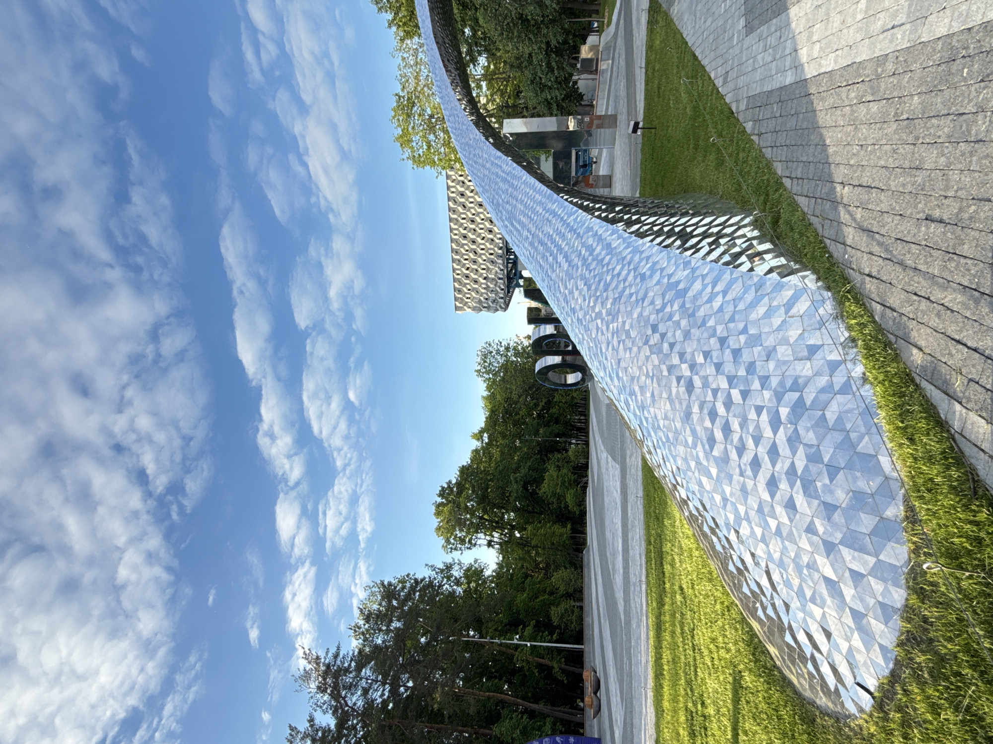 The image captures a vibrant, sunlit scene within a modern urban park, likely Zaryadye Park in Moscow, Russia. The most prominent feature is a long, curved, metallic structure that sweeps from the foreground into the mid-ground. Its surface is composed of numerous small, reflective triangular facets, creating a shimmering, geometric pattern that catches and reflects the blue sky and surrounding greenery.
Adjacent to this structure, a strip of lush green grass runs alongside a wide, textured paved walkway or path, crafted from rectangular stone blocks. In the background, dense clusters of tall, green trees form a natural boundary, suggesting a park or wooded area. Beyond the trees and partially obscured, other contemporary buildings with distinctive architectural styles are visible; one particular structure exhibits a striking hexagonal pattern on its facade.
The sky is a clear, bright blue, dotted with scattered white, wispy clouds, indicating good weather. The strong natural light suggests it is daytime, possibly morning or afternoon. No people are visible in the frame, and there is no apparent activity taking place, giving the scene a serene and architectural focus. There is no visible text in the image.