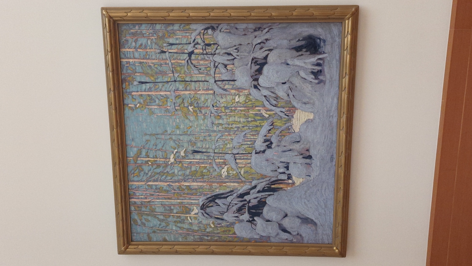 This is a photograph of a framed oil painting depicting a winter forest scene. The painting features tall, slender trees with a pale blue sky visible through their branches. The foreground is dominated by snow-covered trees and bushes, rendered with thick, textured brushstrokes. Small white shapes, possibly birds, are scattered throughout the upper portion of the painting. The painting is in a golden, ornate frame. The photograph is taken from a slightly elevated angle, showing a plain white wall above the artwork and a reddish-brown wooden surface below it. The painting is displayed indoors, likely in a gallery or home. The exact location in Toronto, Canada is not discernible from the image alone.