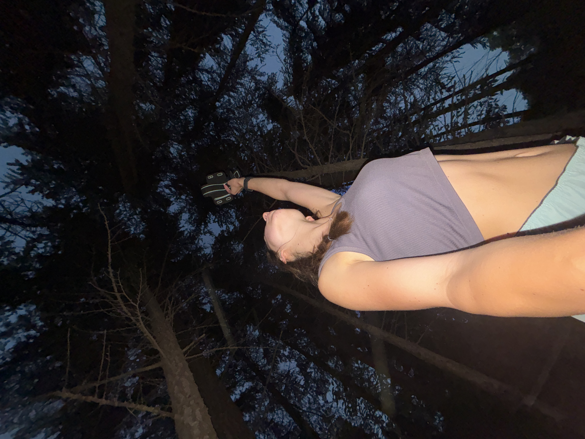 In the city of Varna, Bulgaria, a young woman stands in a forest at dusk. She is viewed from a low angle, looking upwards. She is wearing a purple crop top and light blue shorts. Her right arm is raised, holding a black object with a white strap. The background is filled with the dark silhouettes of tall pine trees, with a sliver of twilight sky visible through the branches. The scene appears to be set in a natural, outdoor environment within Varna.