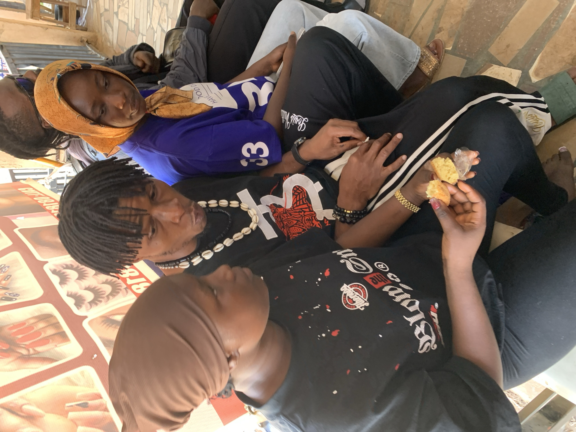 A medium-angle overhead shot depicts a group of people seated closely together on a partially covered stone surface in Jos, Nigeria. The foreground features an advertisement poster for beauty services on the lower left. A person with a light brown headscarf and dark skin is seated, wearing a black t-shirt with a white logo and red accents. Adjacent is a person with dark skin and dreadlocks, wearing a black t-shirt and a beaded necklace. A woman with a gold headscarf and a blue top with white lettering is seated next to them.
Several hands are visible: one pair holds broken pieces of a light-colored baked good wrapped in plastic, and another rests nearby. The attire of others seated includes black pants with white stripes, a blue top with a white number "33", grey denim, and brown sandals. Additional individuals are partially visible, suggesting a crowded setting. The background shows a tiled stone surface.