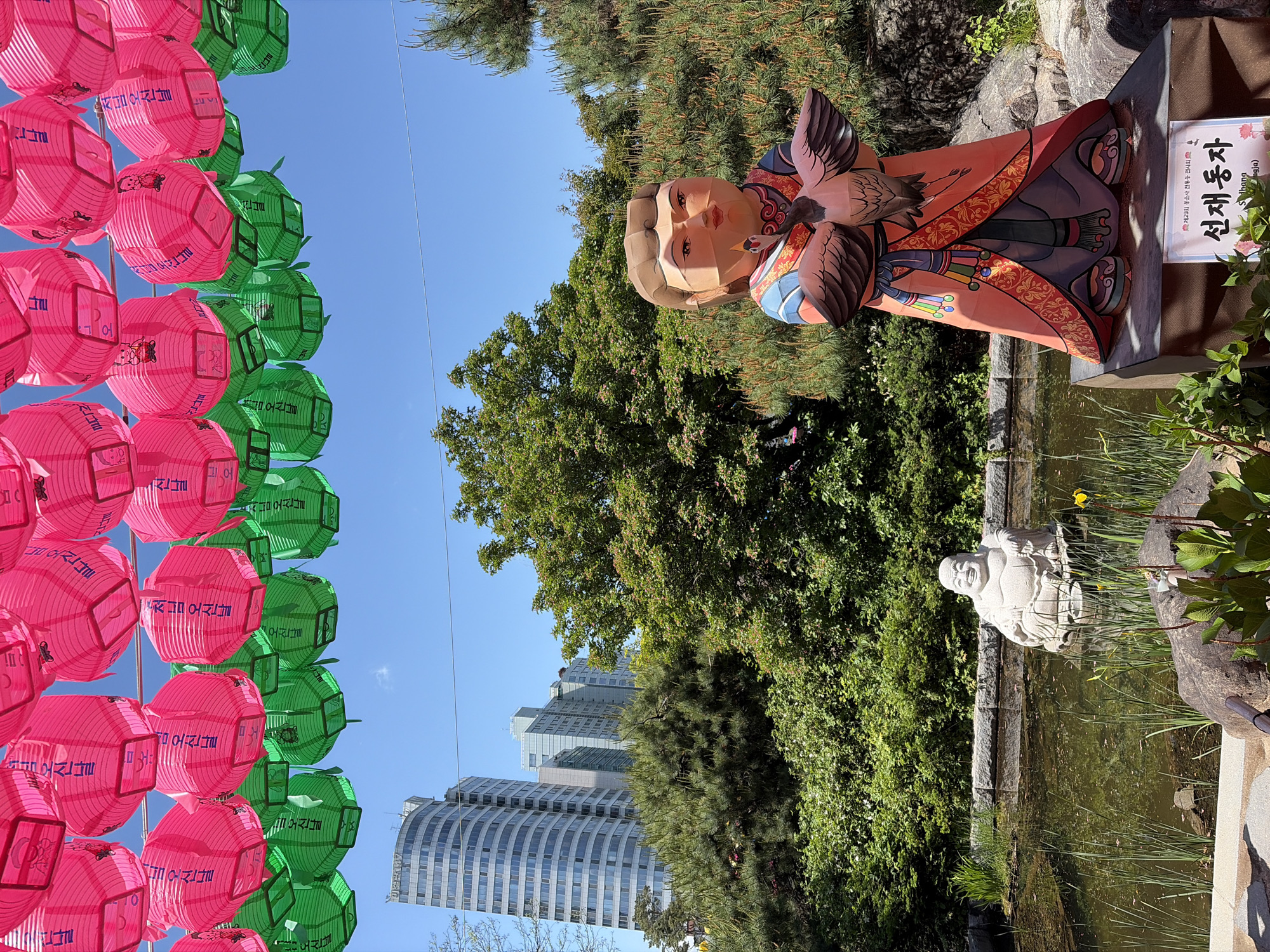 This outdoor shot shows a garden setting with cultural elements in Seoul, South Korea. In the foreground, there are rows of vibrant pink and green lanterns with Korean text. A decorative figure of a person holding a bird stands near a pond with a white statue of a seated figure. Lush greenery surrounds the pond. In the background, tall buildings indicate an urban setting, set against a clear blue sky. A sign with Korean writing is also visible near the decorative figure.
