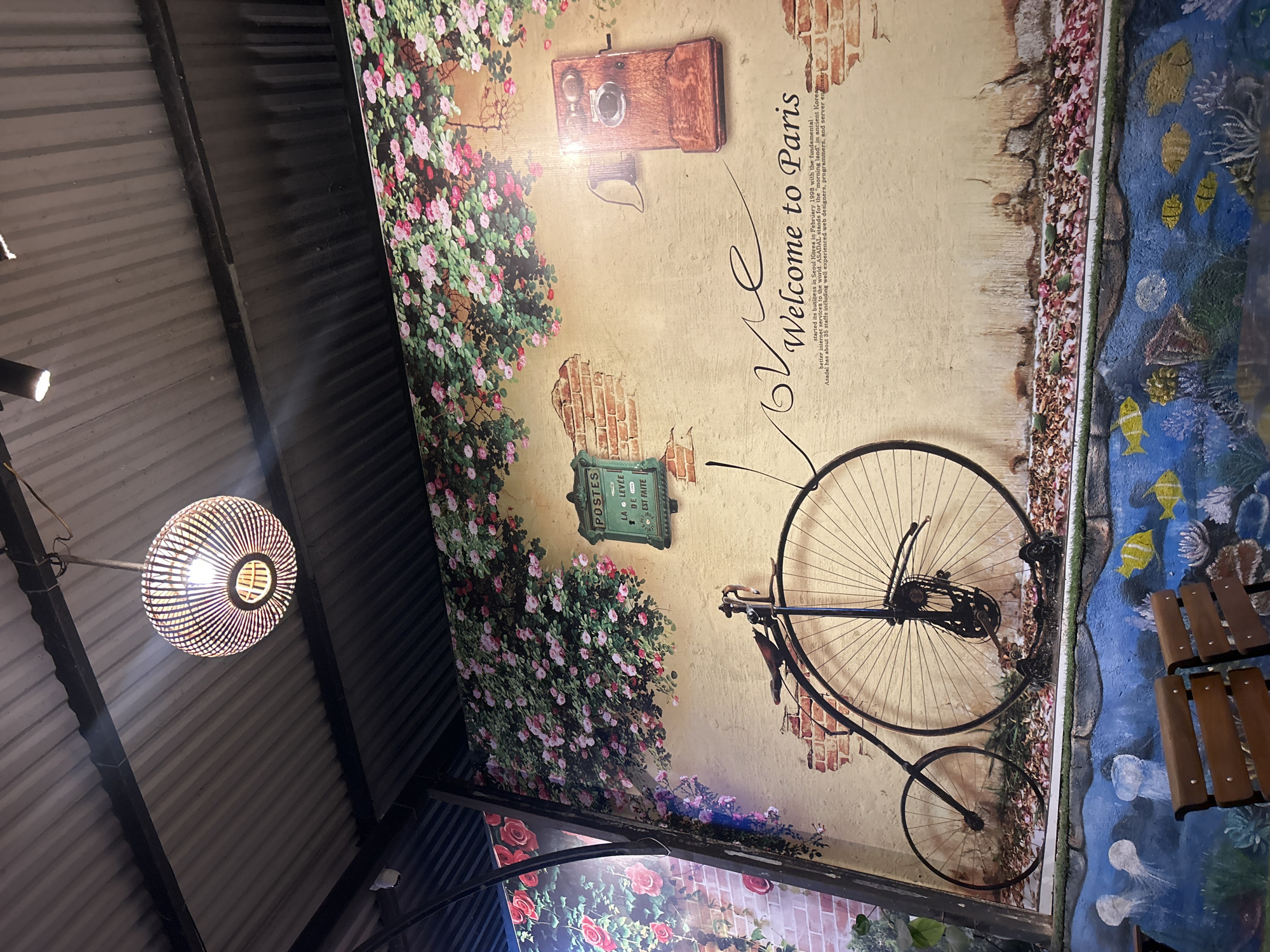The image depicts an interior space in Bắc Nha Trang, Vietnam, featuring a ceiling adorned with themed wallpaper. The wallpaper presents a collage of images related to Paris, including floral arrangements, a vintage camera, a mailbox with "POSTES" inscribed, the phrase "Welcome to Paris," and a bicycle graphic. Adjacent to the Parisian theme is a painted scene resembling an underwater seascape. A light fixture with a woven design hangs prominently from the ceiling, which appears to be corrugated metal. A table with wooden slats is partially visible. The overall perspective is angled upwards towards the ceiling.