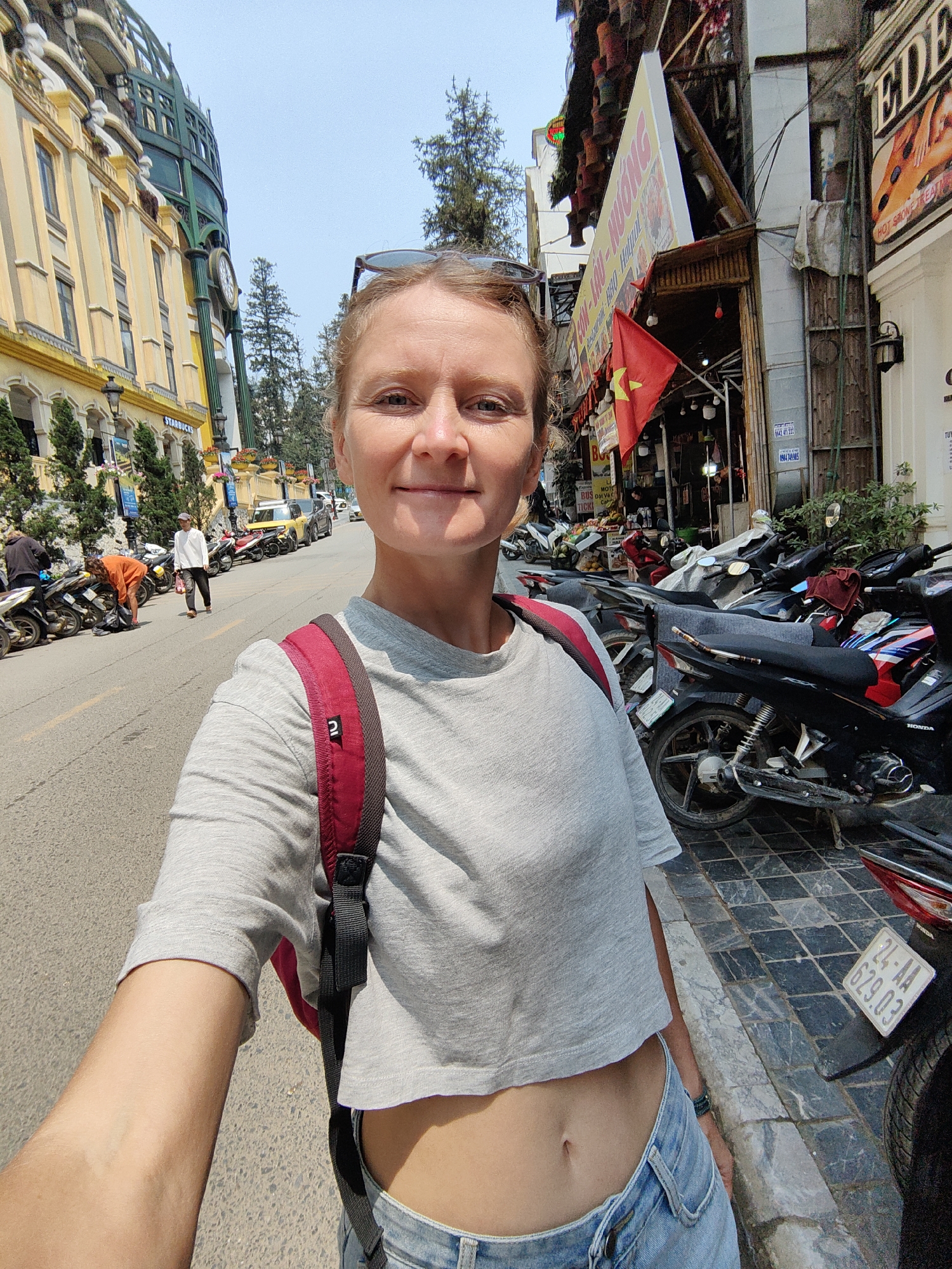 A fair-skinned woman with light brown hair pulled back, wearing sunglasses atop her head, is captured in a front-facing selfie on a sunny day. She wears a grey cropped t-shirt, light blue jeans, and a red backpack with black straps. Her midriff is exposed. She holds the camera with her right arm, extended forward and slightly to the left.
The scene is an urban street in Sa Pa, Vietnam. To her left, the road is lined with numerous parked motorbikes, primarily scooters. Further down the street, several individuals are visible: one person in a white shirt and dark pants walks away from the camera, another in an orange shirt is bent over near a motorbike, and a third in a dark hoodie stands further back. A light yellow multi-story building with ornate facades and a prominent dark green domed or rounded structure featuring a clock face stands on the far left. A "Starbucks" sign is visible at its base.
To her right, a tiled sidewalk with dark grey and black patterns runs alongside a row of businesses. A red Vietnamese flag with a yellow star is displayed prominently from one establishment. Signs with Vietnamese text, including "CƠM - NƯỚNG" and another for "EDEN Hot Stone Treatments," are visible. More motorbikes are parked on this sidewalk and along the street edge, one displaying a Vietnamese license plate "24-AA 629.03." Tall, slender trees are visible in the background, suggesting a surrounding natural landscape. The sky is clear and bright.