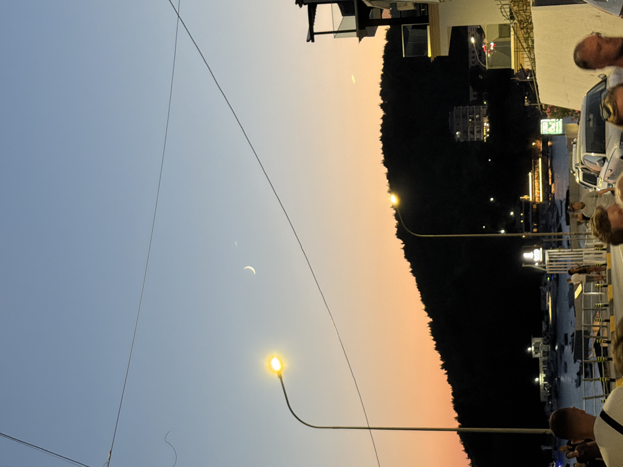 The image captures a twilight scene in Himarë, Albania, looking towards the coastline. A crescent moon is visible in the pale blue sky, transitioning to an orange hue near the horizon. A silhouetted hillside with trees dominates the midground, while the foreground features a waterfront promenade with people, parked cars, and buildings. Streetlights illuminate the scene, casting a warm glow. Several boats are moored in the bay. The overall atmosphere is serene, indicative of a peaceful evening by the sea.