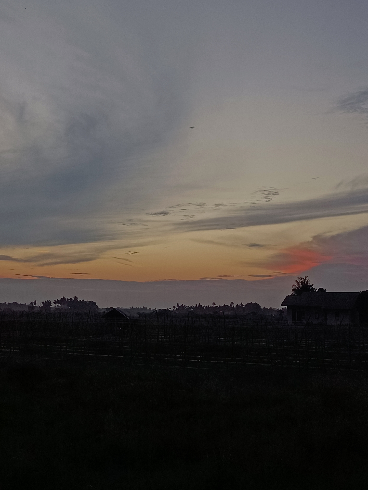 The image captures a twilight sky over a rural landscape in Meunasah Pupu, Indonesia. The sky is a gradient of muted blues, grays, and oranges, with streaks of pink near the horizon. Wispy clouds stretch across the sky. In the distance, a silhouette of trees and vegetation is visible. A small, dark-colored building with a visible roofline is situated on the right side of the frame, and another smaller structure is partially visible to the left of it, amidst the distant treeline. The foreground is a dark silhouette, obscuring details of the land and any potential inhabitants or ongoing activities. A faint, dark linear object, possibly a distant airplane, is visible in the upper left quadrant of the sky. The scene appears serene and still, indicative of either dawn or dusk.