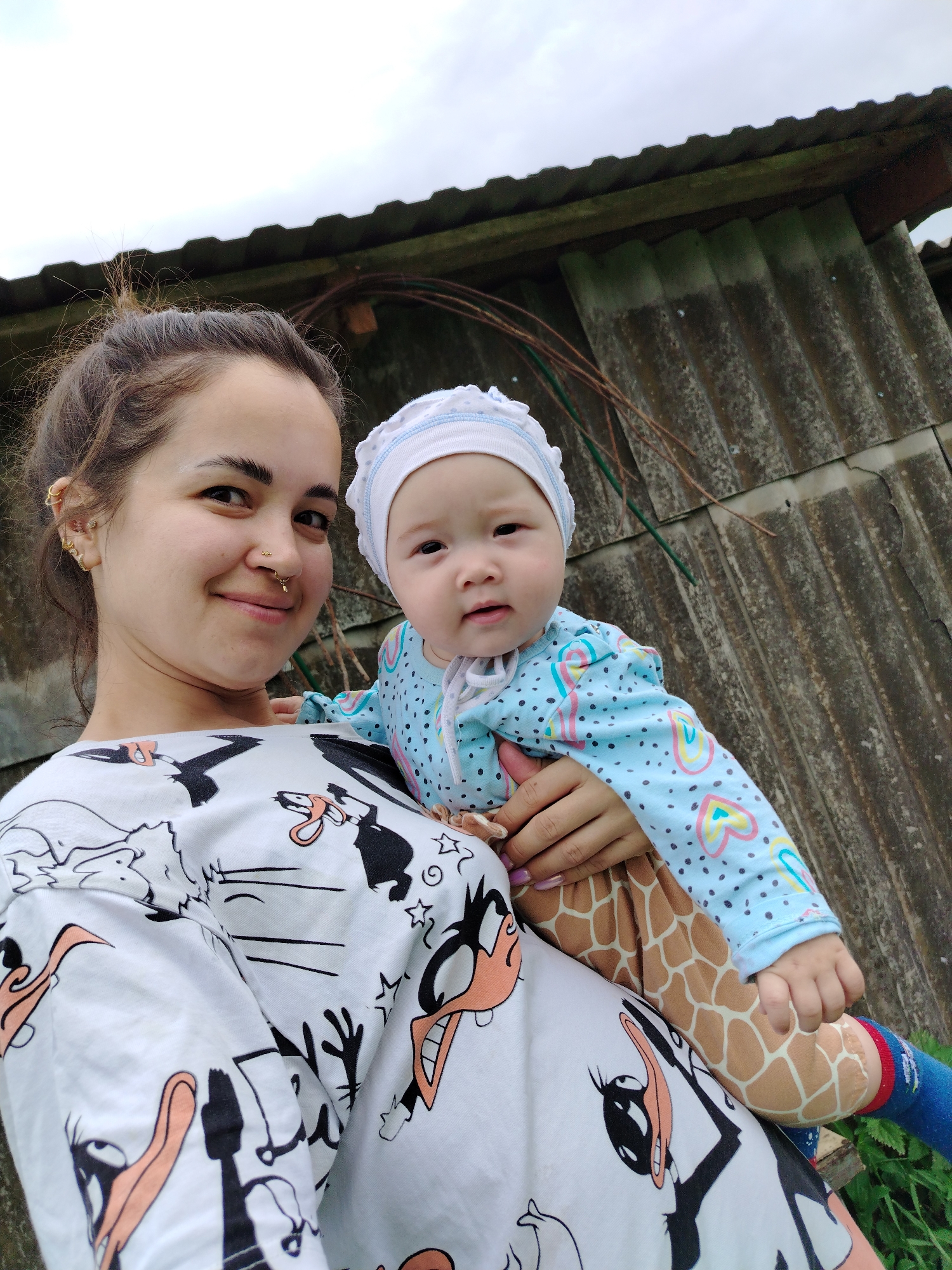A young woman is holding a baby in front of a rustic, weathered corrugated metal wall. The woman has dark hair pulled back and is wearing a white shirt with a cartoon character print, featuring black and orange figures with large beaks and eyes. She is smiling at the camera. The baby is wearing a light blue long-sleeved outfit with colorful heart patterns and polka dots, and a white bonnet with a ruffled edge. The baby is looking towards the camera with an inquisitive expression. The baby's legs are clad in what appear to be giraffe-patterned leggings and blue socks with red and white stripes. The scene appears to be outdoors, with overcast skies visible in the background and hints of green foliage at the bottom right. The overall impression is one of warmth and affection between the woman and child, set against a simple, rural backdrop, likely in Гожан, Russia. The lighting suggests it is daytime, possibly late afternoon or early morning given the diffused light.